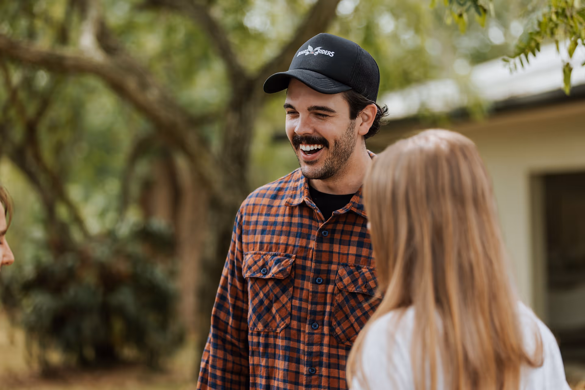 Smiling man in a black cap and orange plaid shirt talking outdoors with two people.