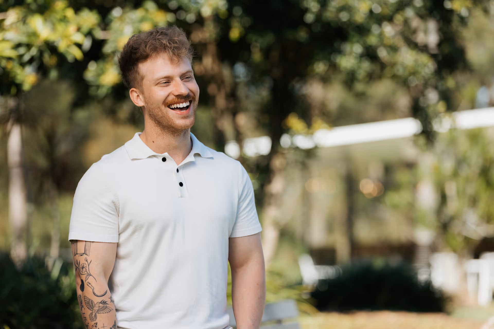Smiling young man with curly hair and tattoos on his left arm wearing a white polo shirt outdoors.