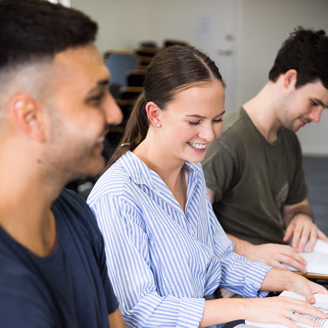 Three students sitting in a classroom reading and smiling, with the central female student wearing a striped shirt.