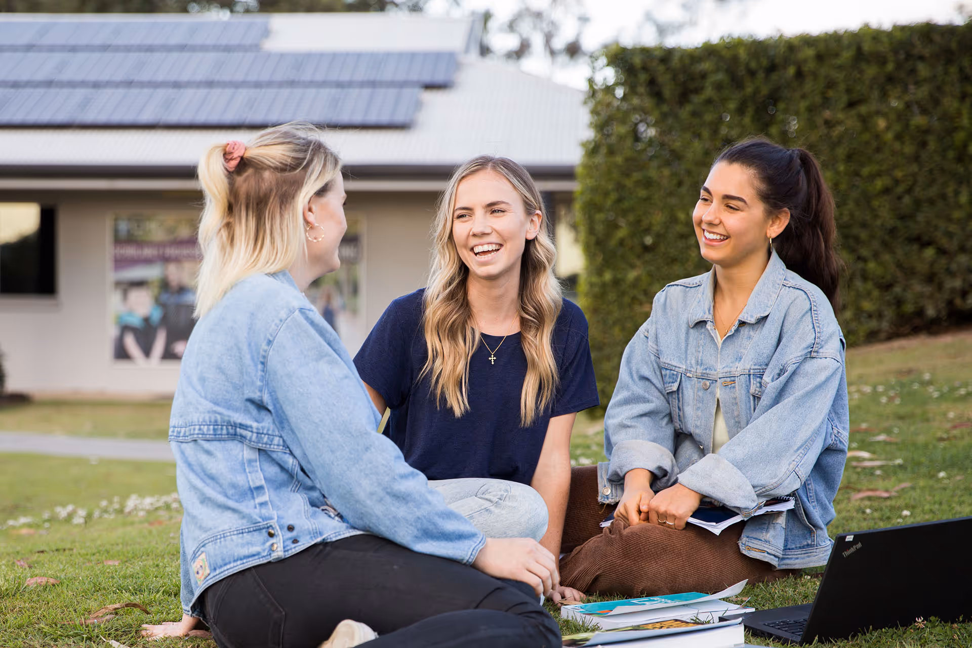 Three young women sitting on grass outside, smiling and talking with notebooks and a laptop nearby.