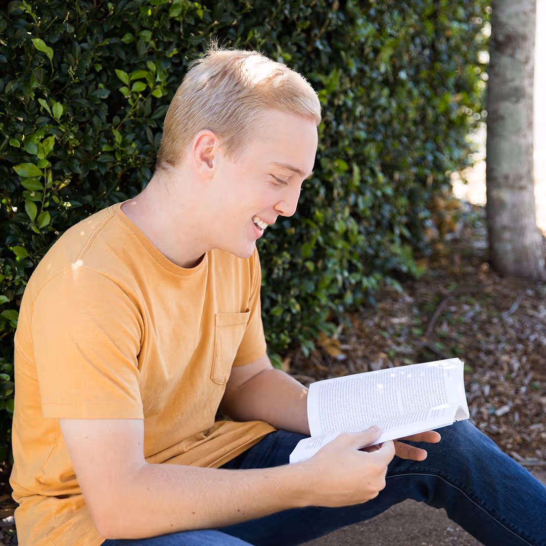 Young man in an orange shirt sitting outdoors reading a book and smiling.