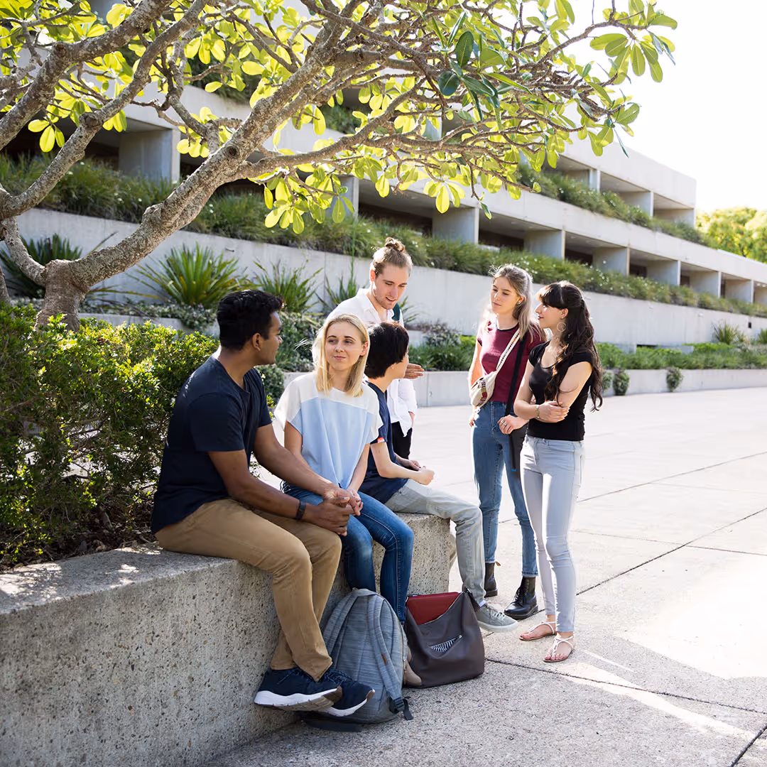 Group of five young adults sitting and standing outdoors near a concrete wall with greenery and a modern building in the background.