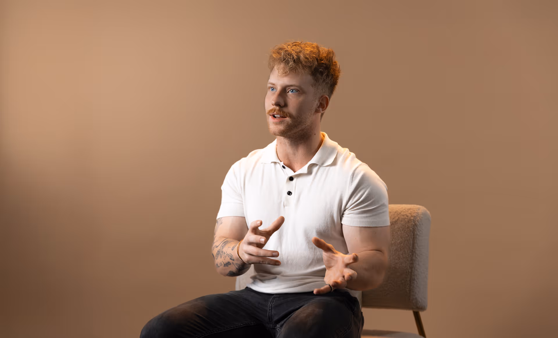 Young man with curly hair and tattoos, wearing a white polo shirt, seated on a chair gesturing with hands while speaking.