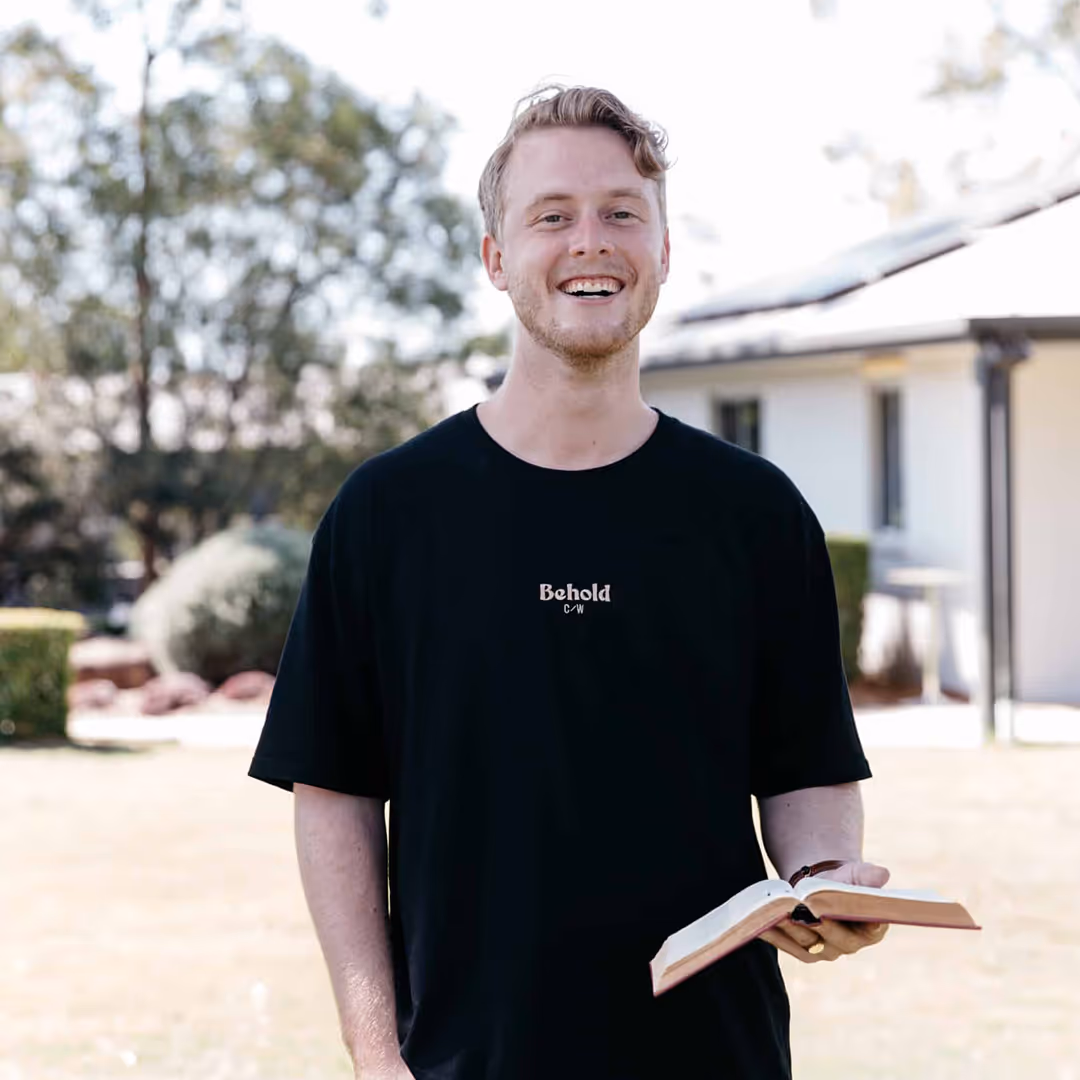 Smiling young man wearing a black t-shirt holding an open book outdoors in a sunny yard.