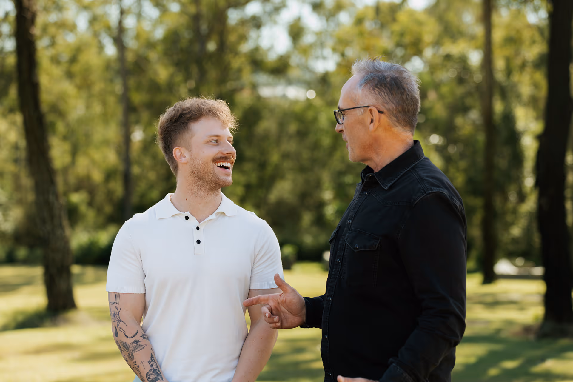 Two men outdoors, one younger with tattoos wearing a white polo shirt smiling, and one older with glasses wearing a black shirt talking.
