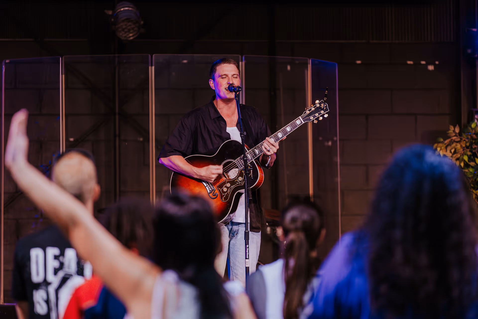 Man playing acoustic guitar and singing on stage in front of an audience with raised hands.