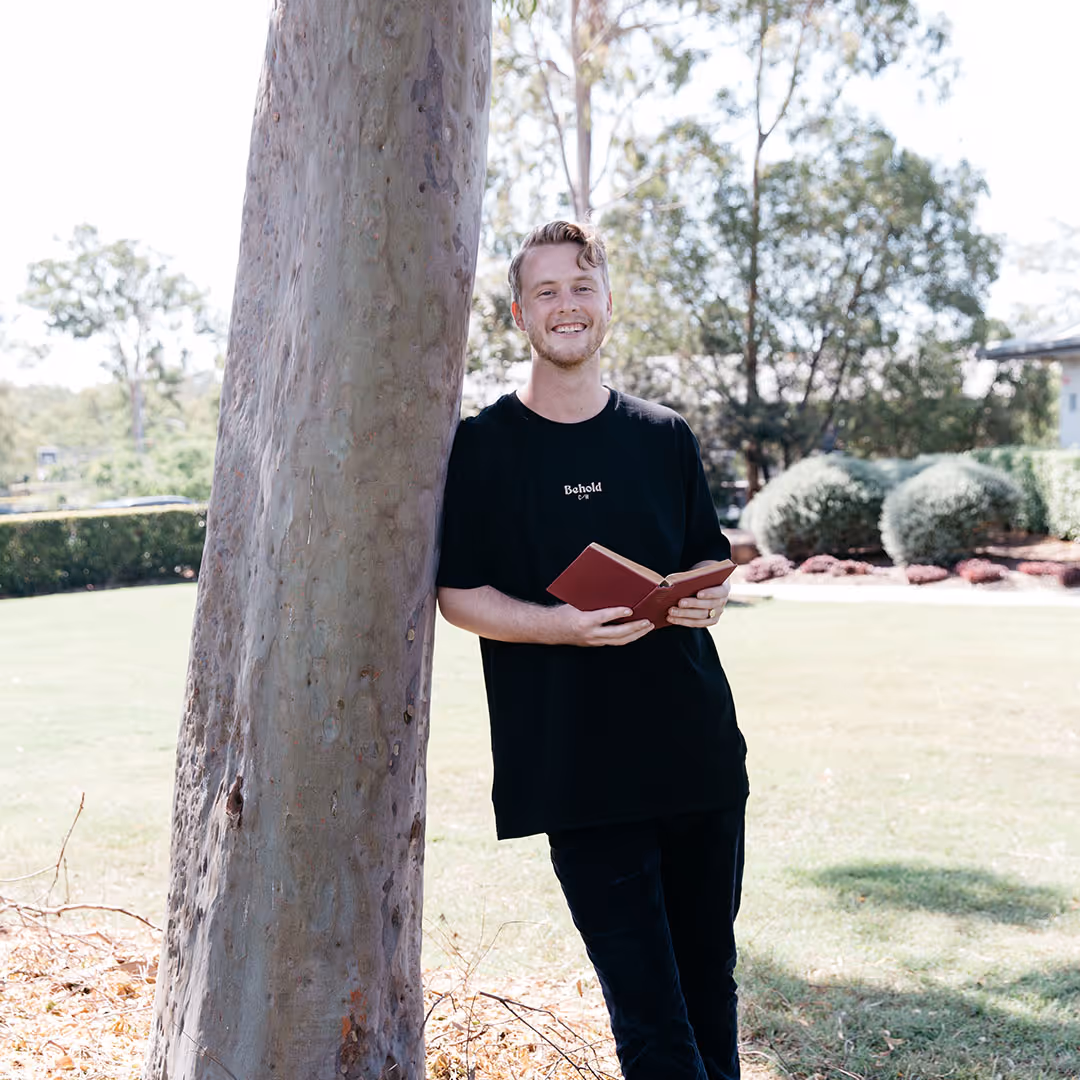 Smiling young man in a black shirt leaning against a tree while holding an open book in a sunny park.