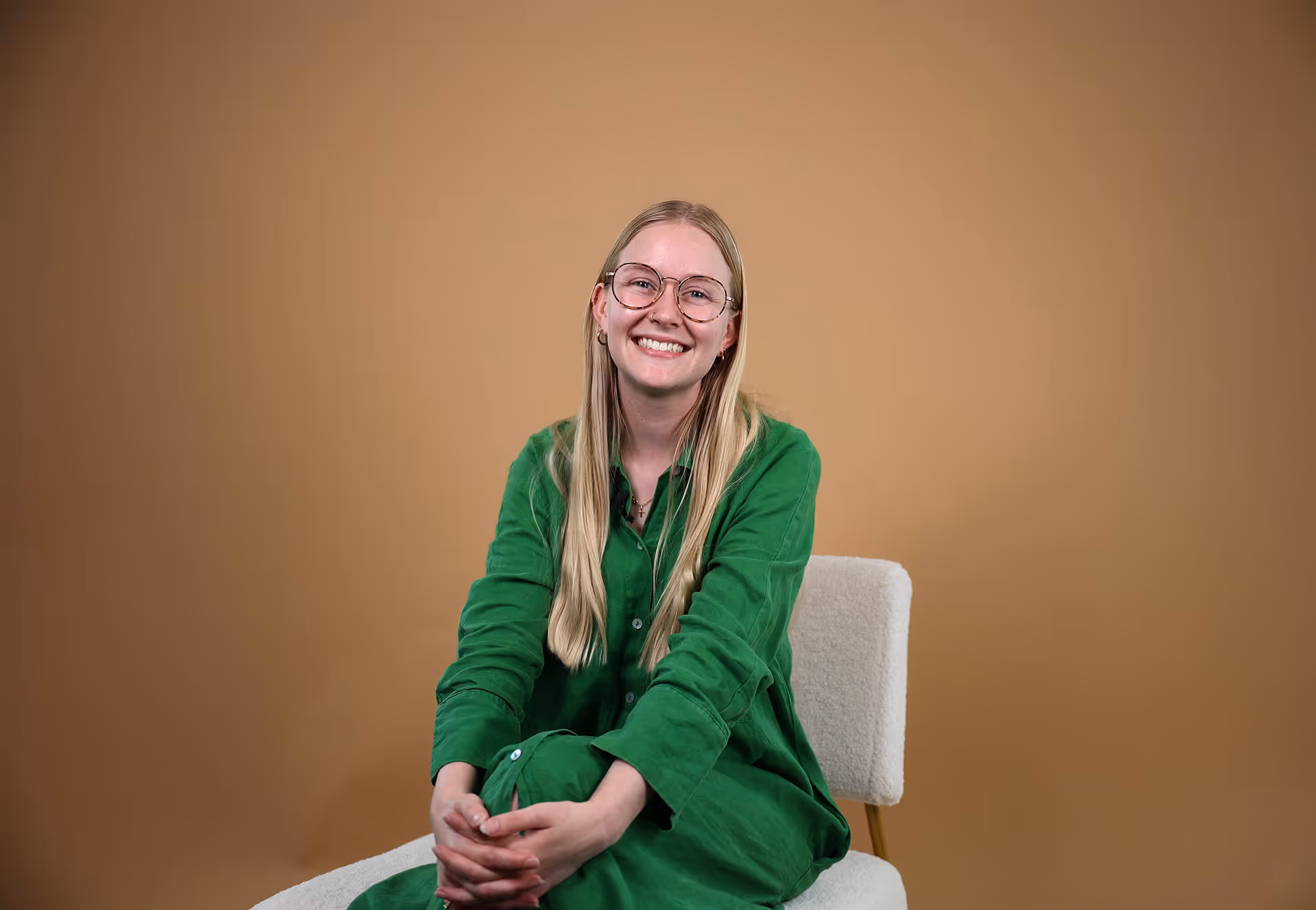 Smiling woman with long blonde hair and glasses wearing a green dress sitting on a white chair against a brown background.