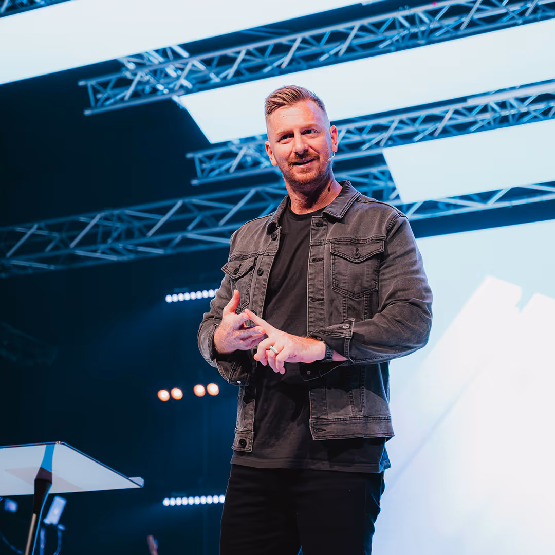 Man with short hair and beard speaking on stage, wearing a dark jacket and black shirt under bright stage lights.