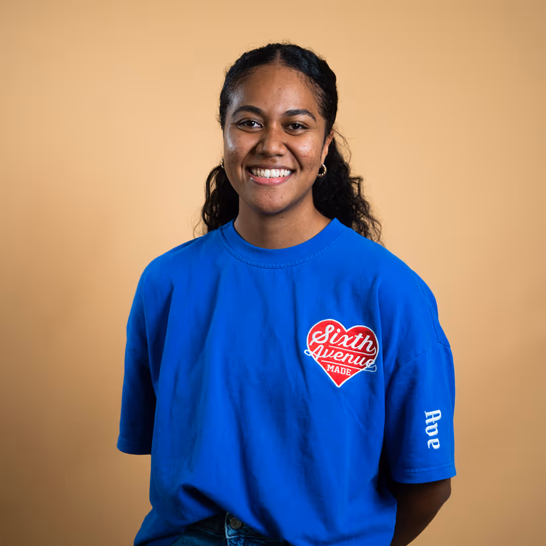 Smiling woman with curly hair wearing a blue shirt with a red heart logo and text on beige background.