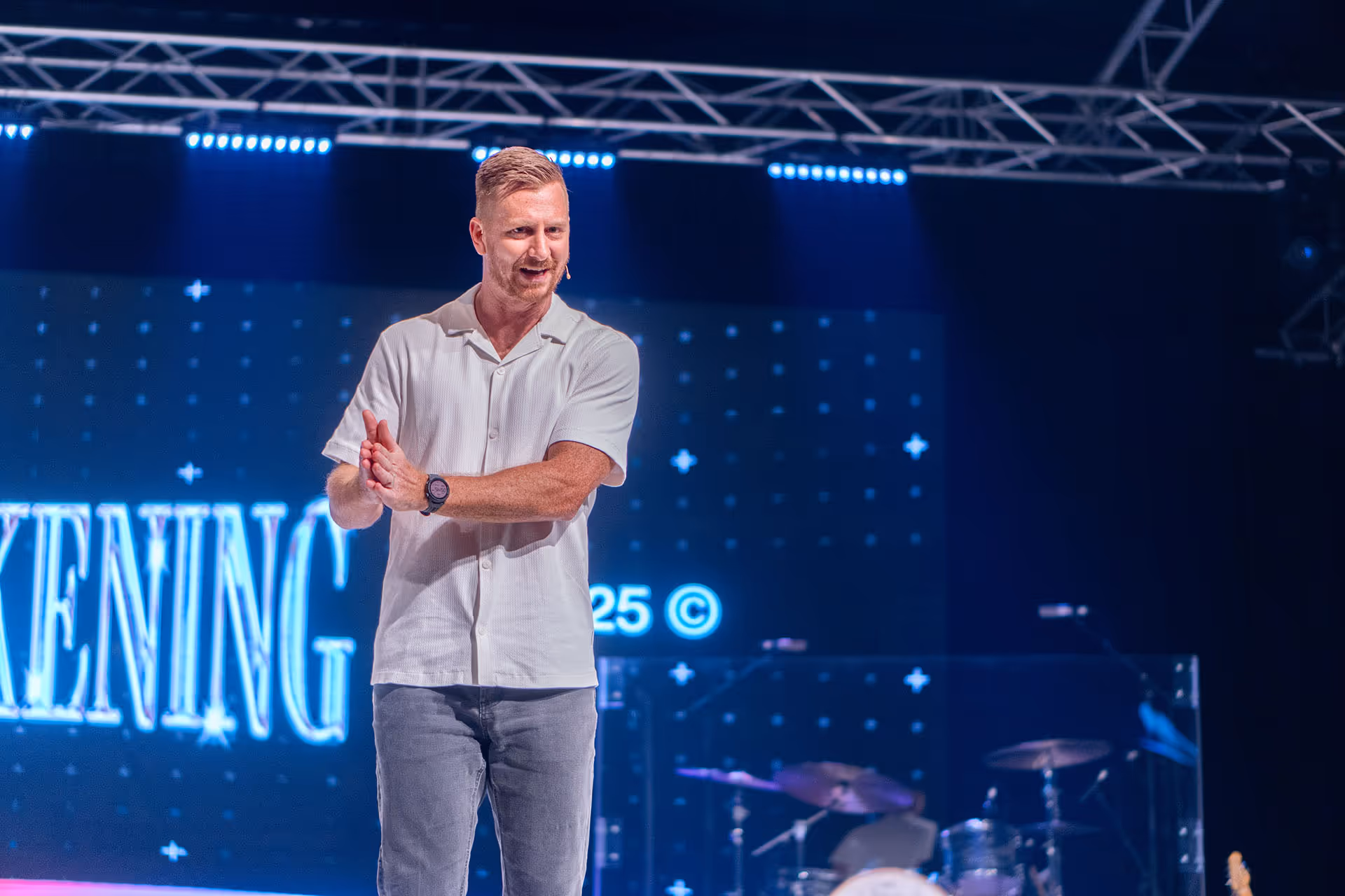 Man speaking on stage with microphone headset, wearing a white short-sleeve shirt and jeans, with a drum set and blue stage lights in the background.