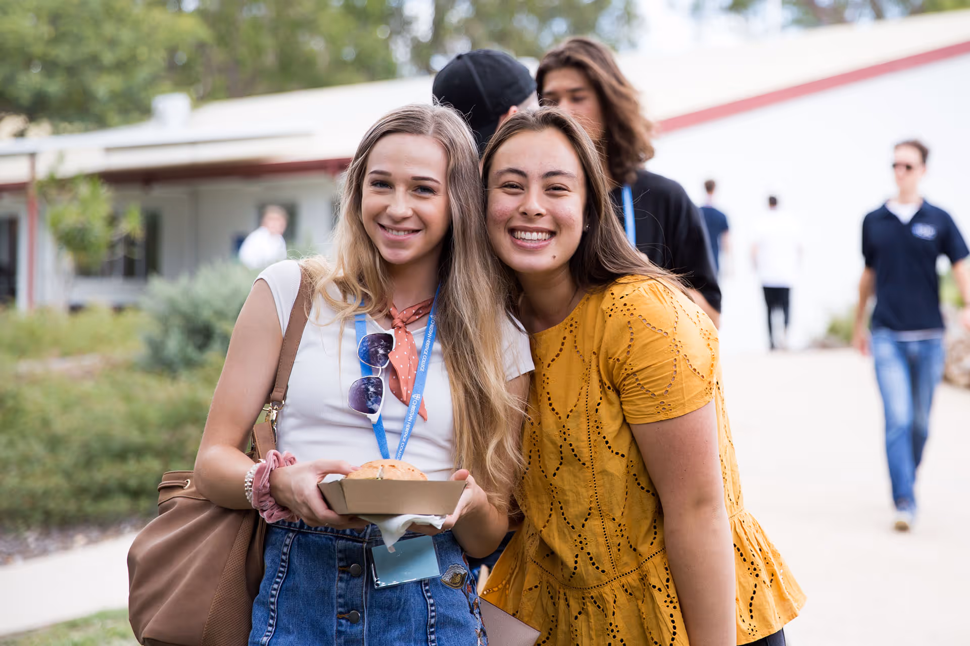 Two young women smiling outdoors, one holding a sandwich in a paper tray, with blurred people and greenery in the background.