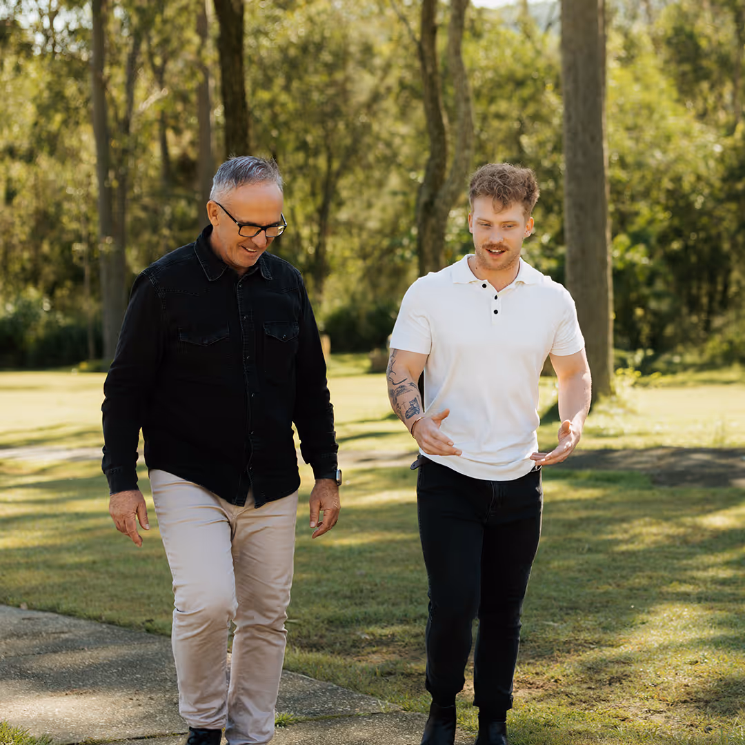 Two men walking and talking outdoors on a sunny day in a park-like setting.