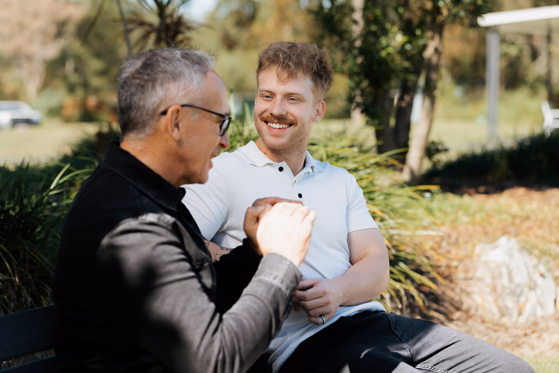Two men sitting outdoors on a bench, smiling and engaged in conversation, with greenery in the background.