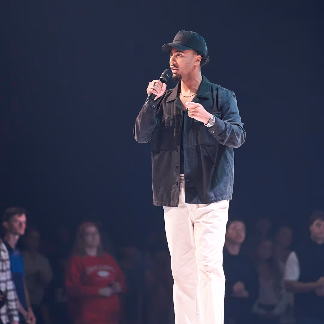 Man wearing a black jacket and cap speaking into a microphone on stage with audience in the background.
