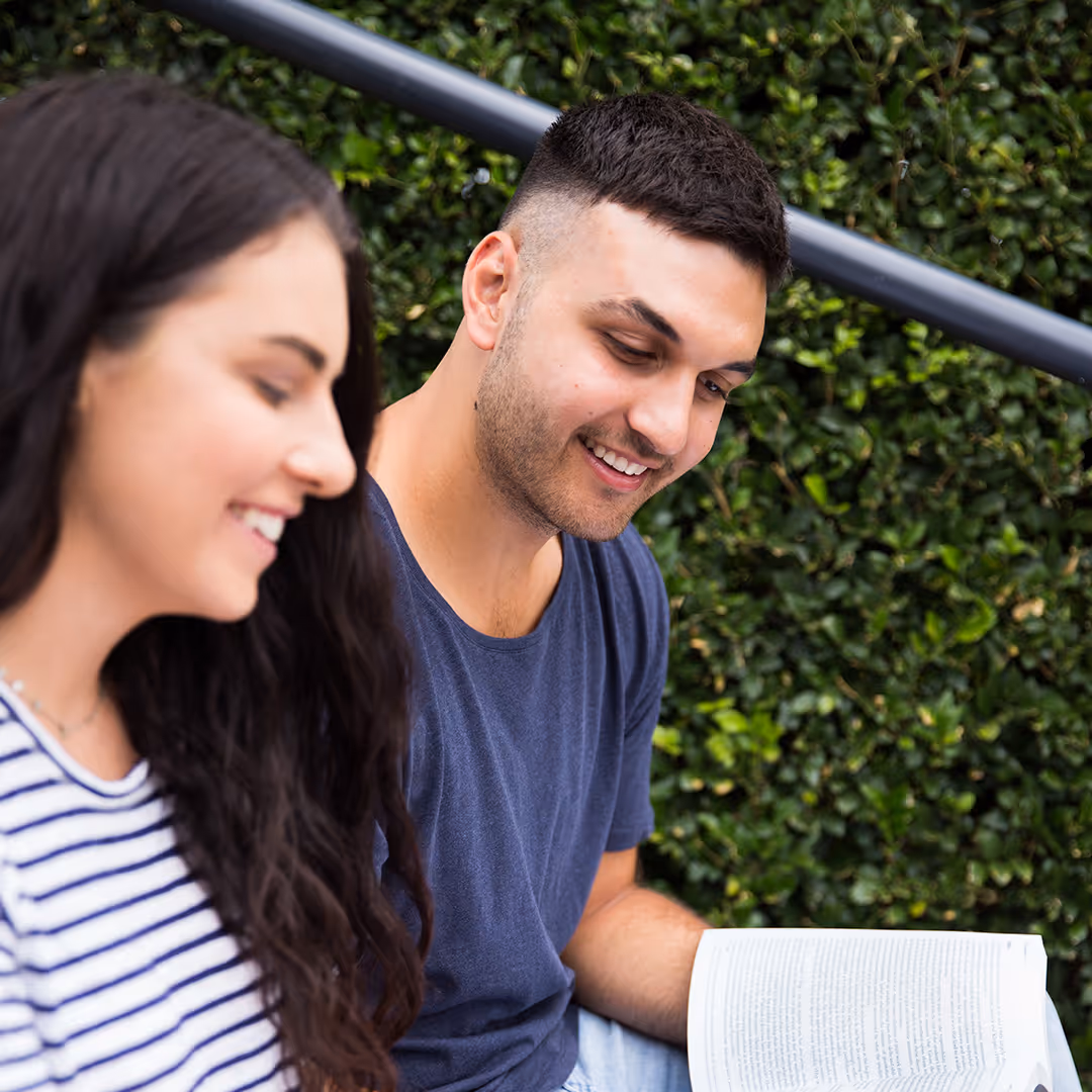 Two young adults sitting outside, smiling and reading a book together.