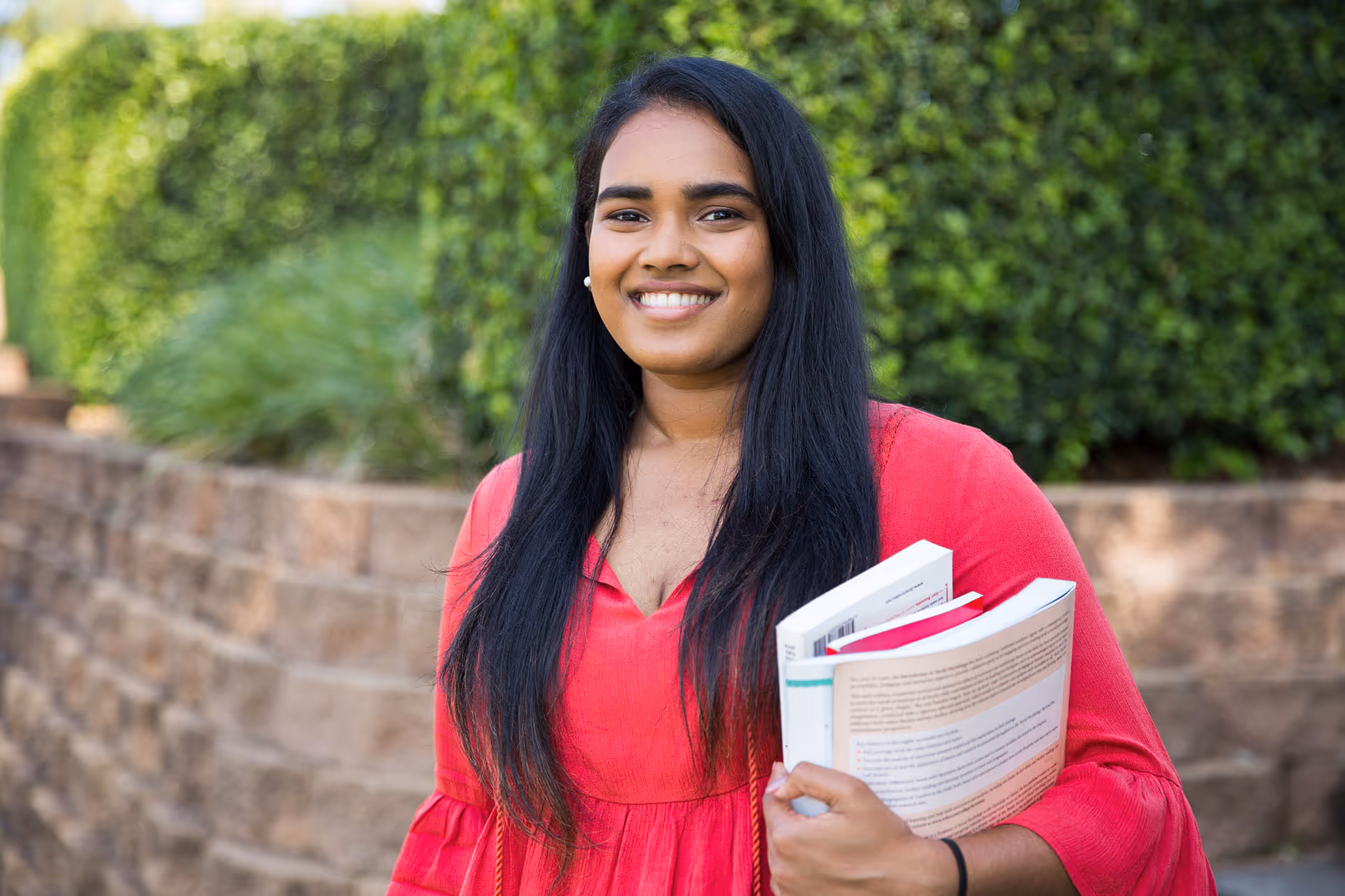Young woman with long black hair wearing a red dress holding books outdoors in front of a stone wall and greenery.