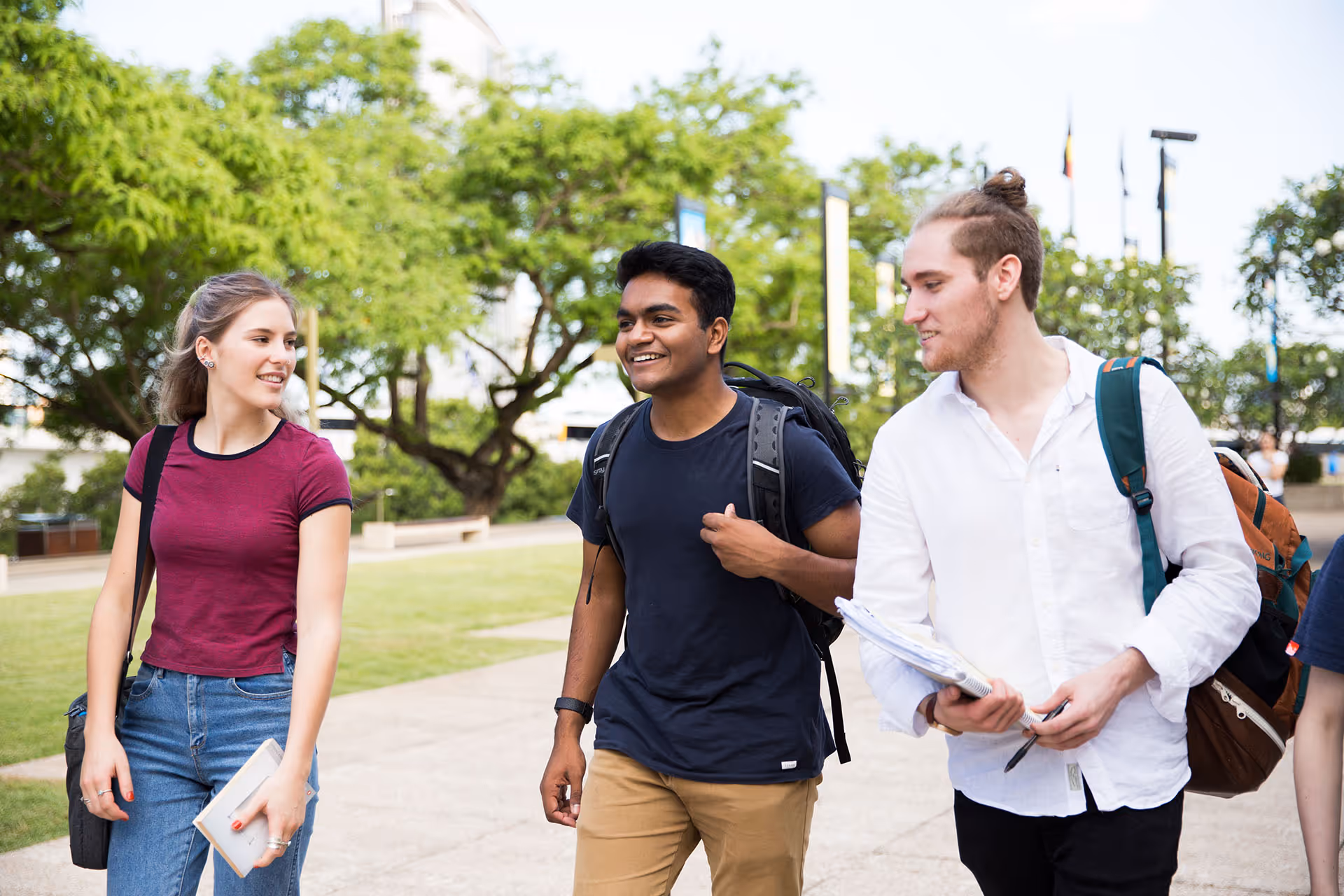 Three college students walking outdoors on campus, smiling and carrying backpacks and notebooks.
