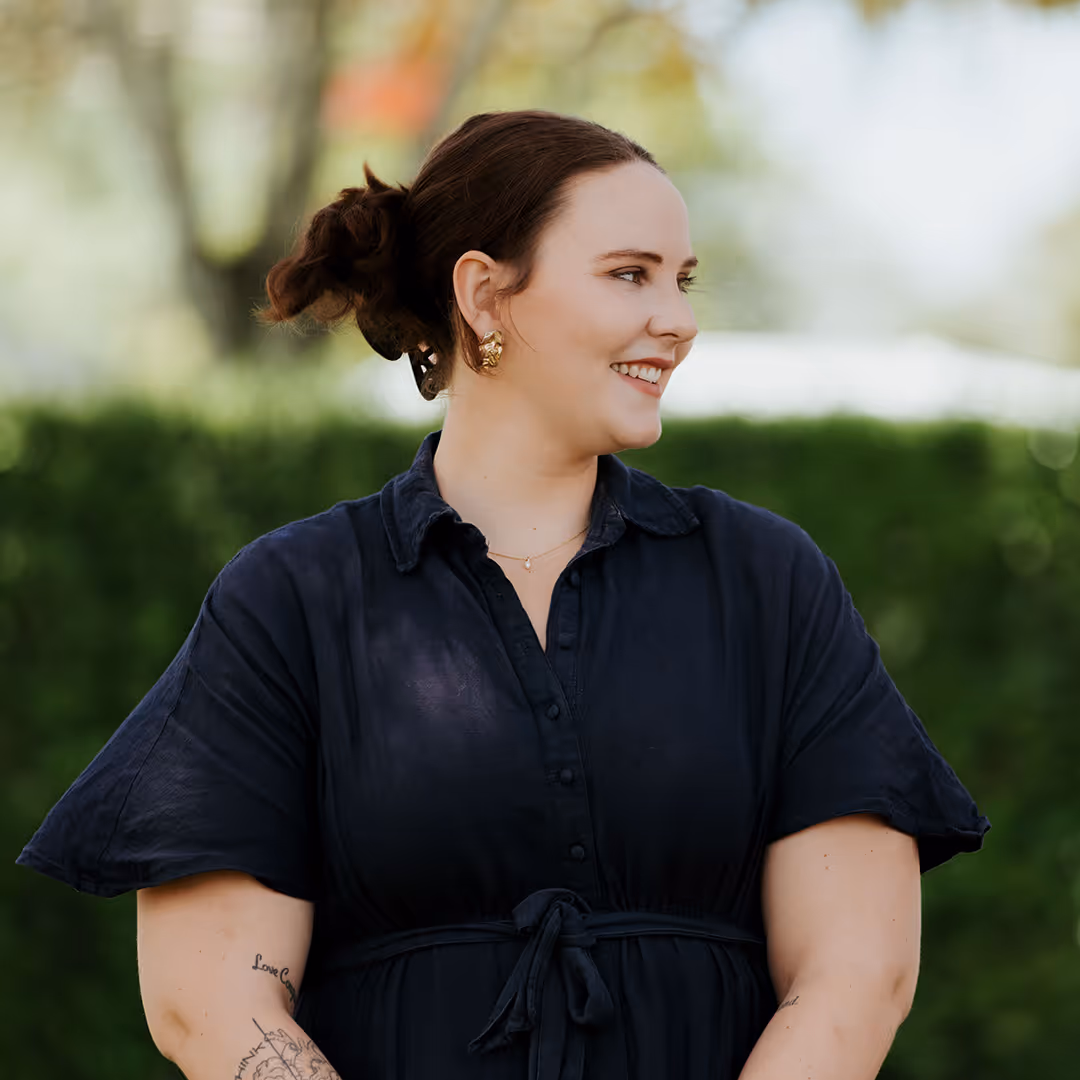 Smiling woman with dark hair tied up, wearing a black dress with short sleeves and gold earrings, standing outdoors with blurred greenery in the background.