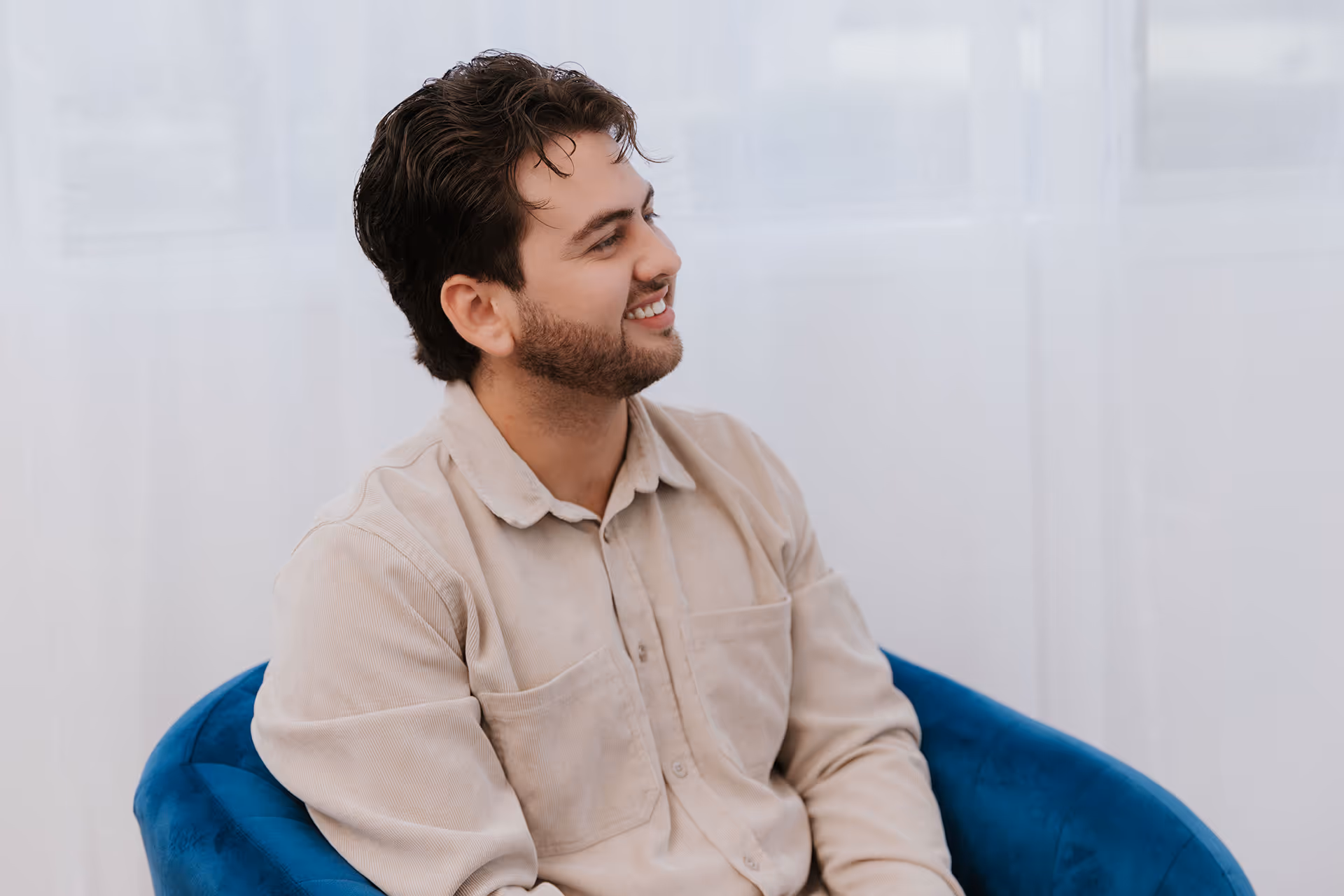 Smiling man with dark hair and beard sitting in a blue chair wearing a beige shirt.