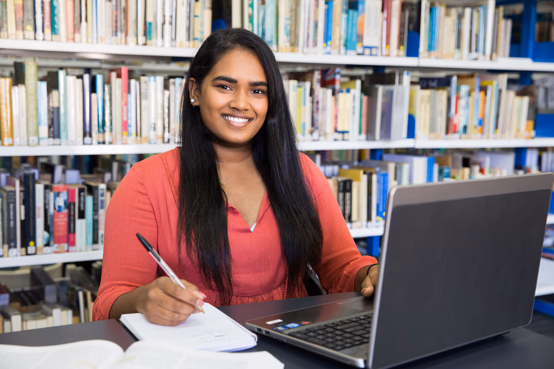 Smiling woman with long dark hair sitting at a table in a library, writing in a notebook with a laptop open in front of her.