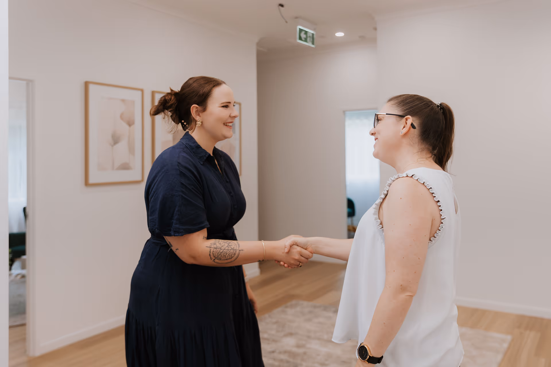 Two women smiling and shaking hands in a bright, modern room with wooden flooring.