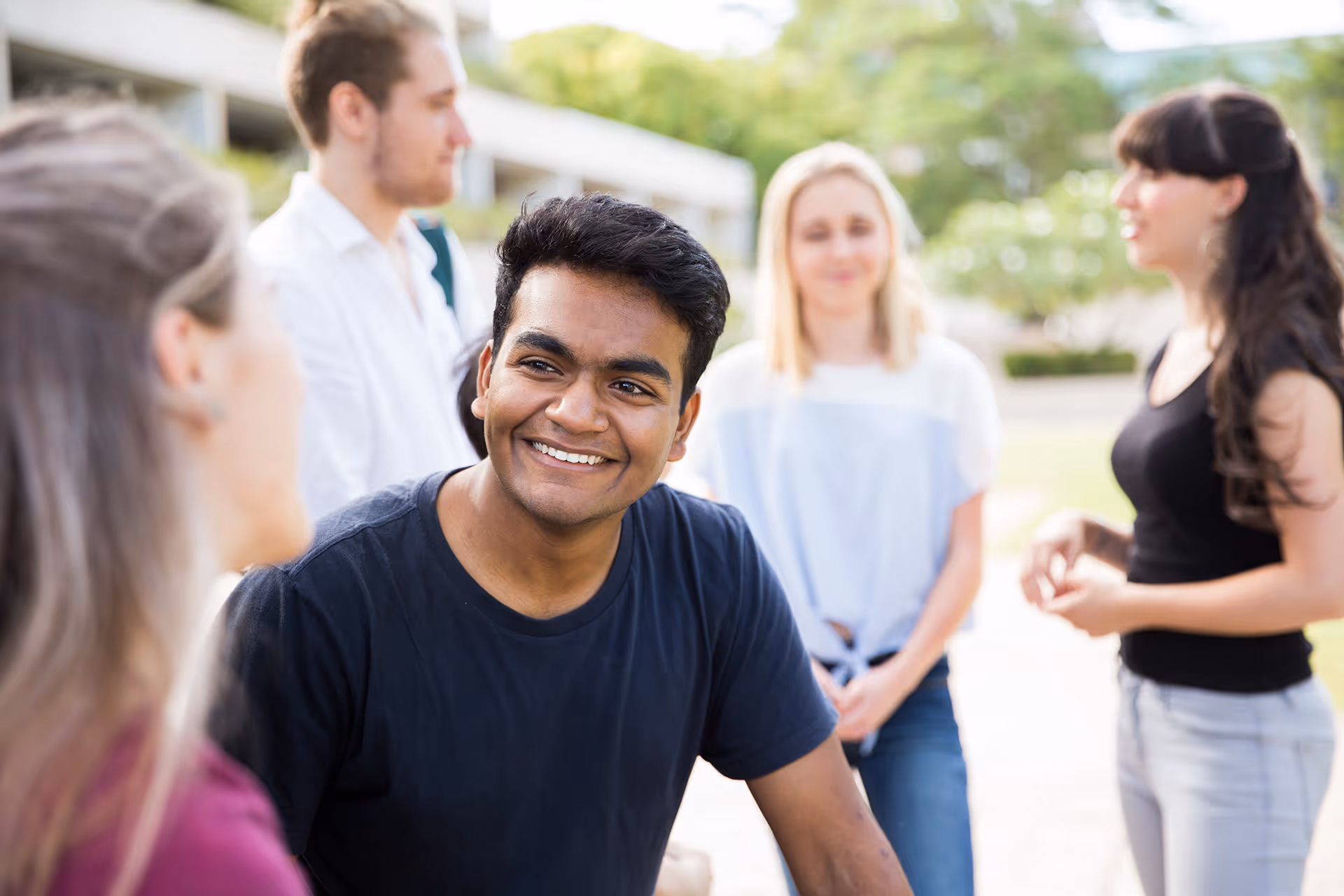 Smiling young man in a black t-shirt talking to a group of four people outdoors.