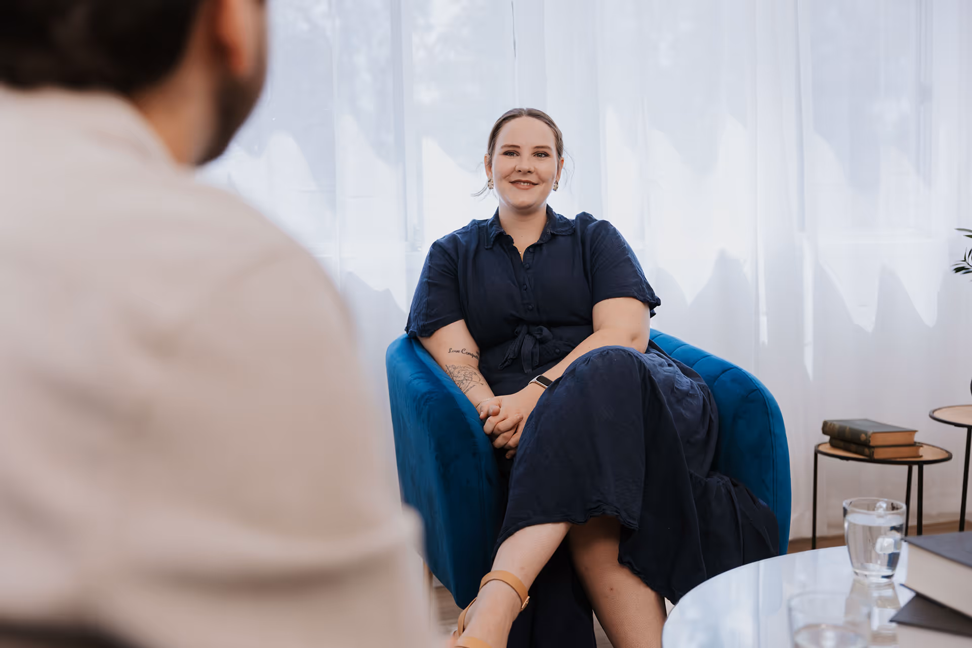 Woman in a navy blue dress sitting cross-legged in a blue chair, smiling during a conversation with a man partially visible in the foreground.