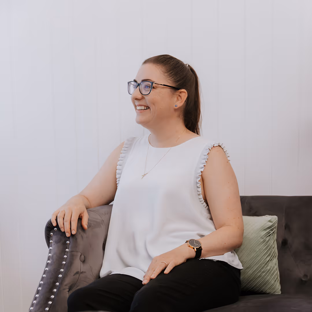 Smiling woman with glasses and a ponytail sitting on a grey couch with a green pillow, wearing a white sleeveless top and black pants.