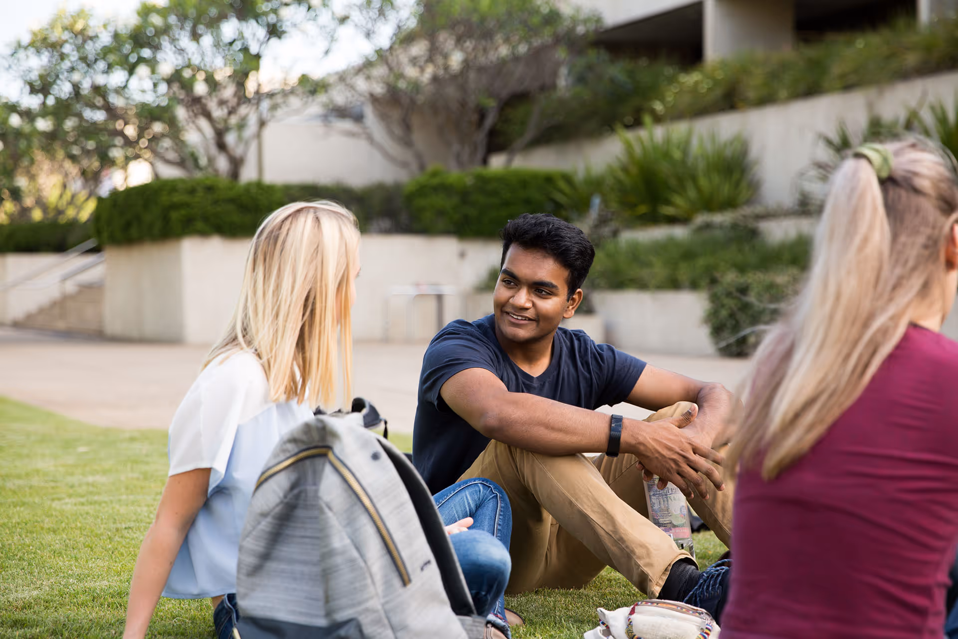 Three young adults sitting on grass outside, engaging in conversation with backpacks and casual clothing.