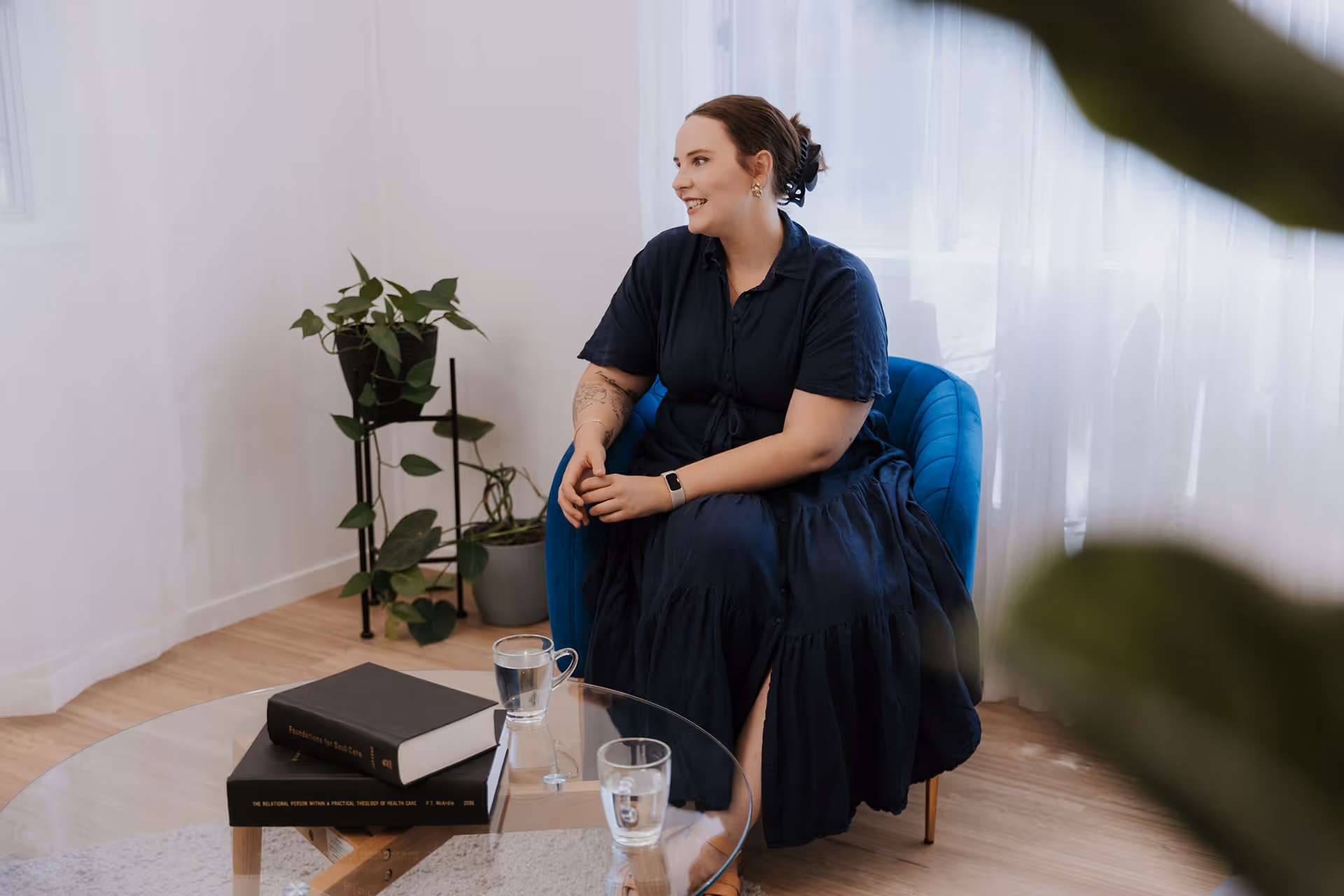 Woman in a dark blue dress sitting on a blue chair smiling inside a room with plants and a glass table with books and glasses of water.