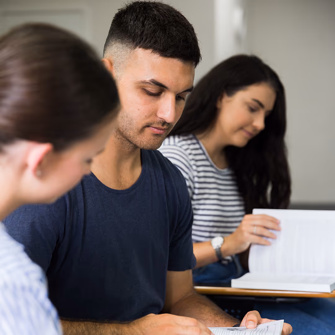 Three students sitting at desks reading and studying books in a classroom.