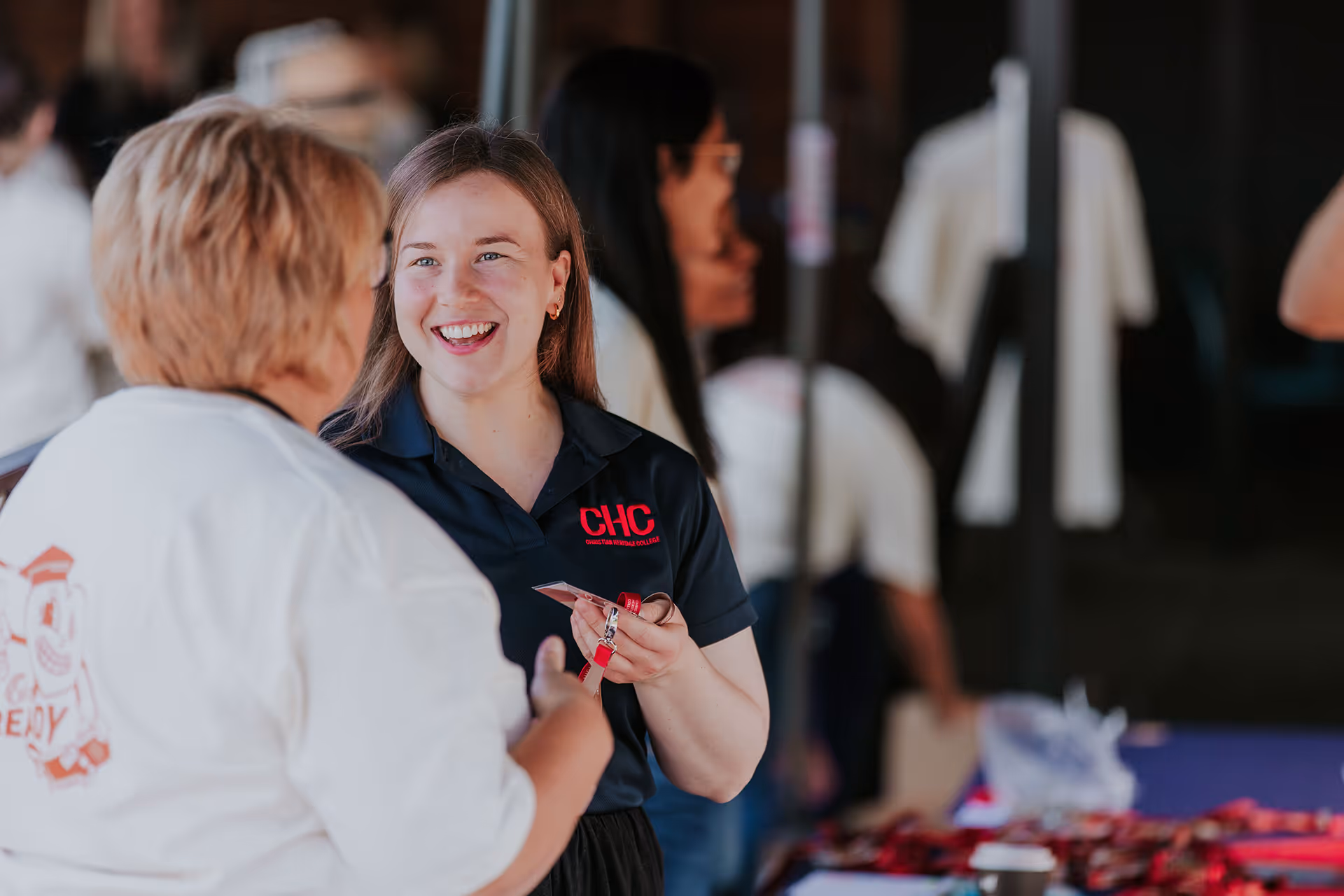Smiling woman in a navy CHC polo engaging in conversation at an event with another person.