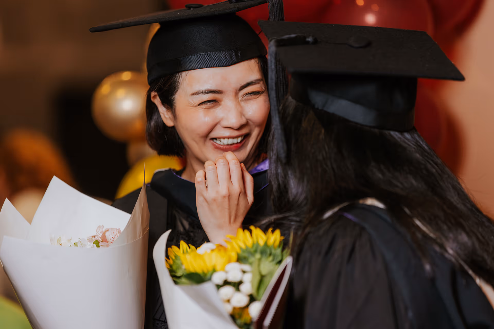 Two graduates in caps and gowns smiling and holding flower bouquets during a celebration.