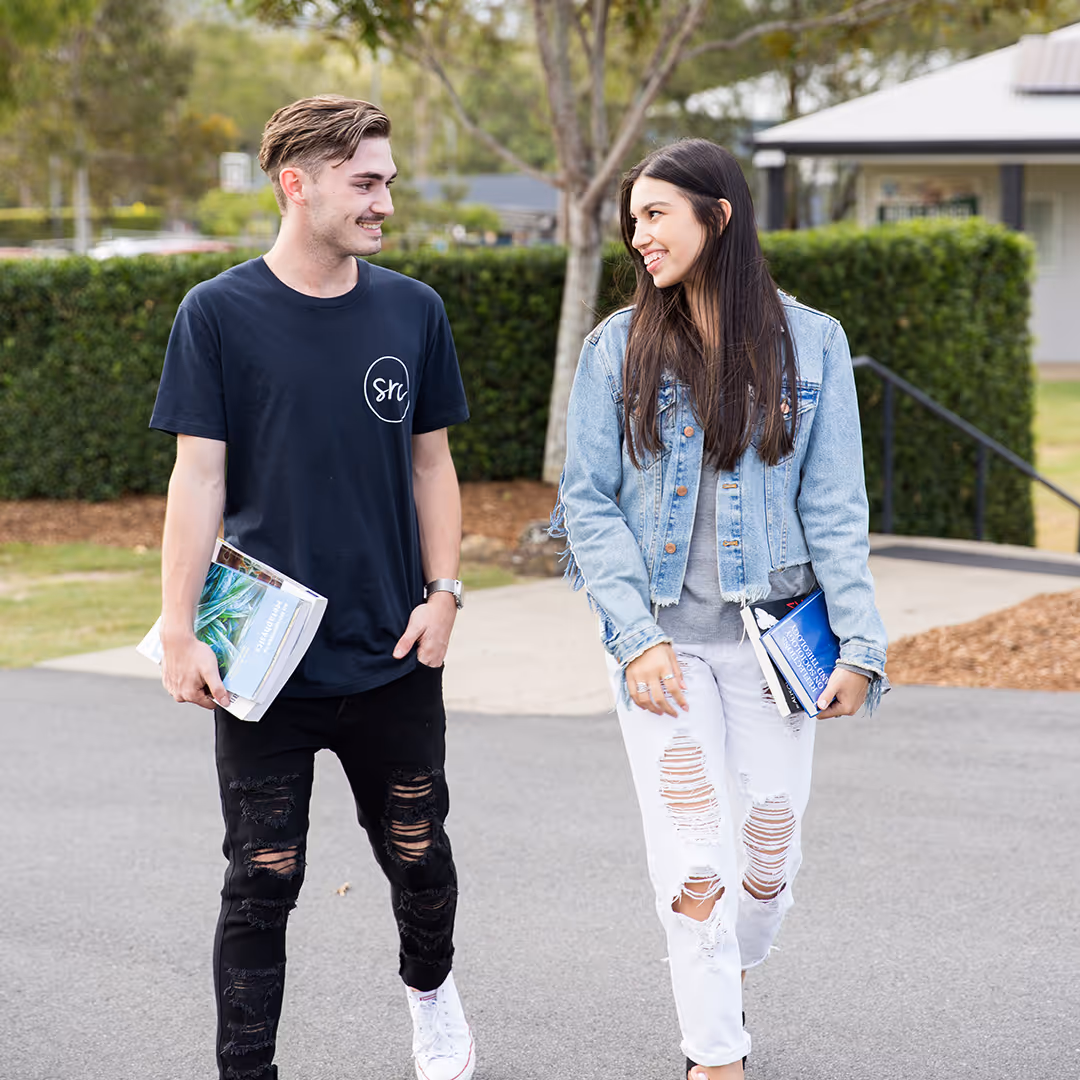 Two young adults walking outdoors on a path, smiling at each other and carrying books.