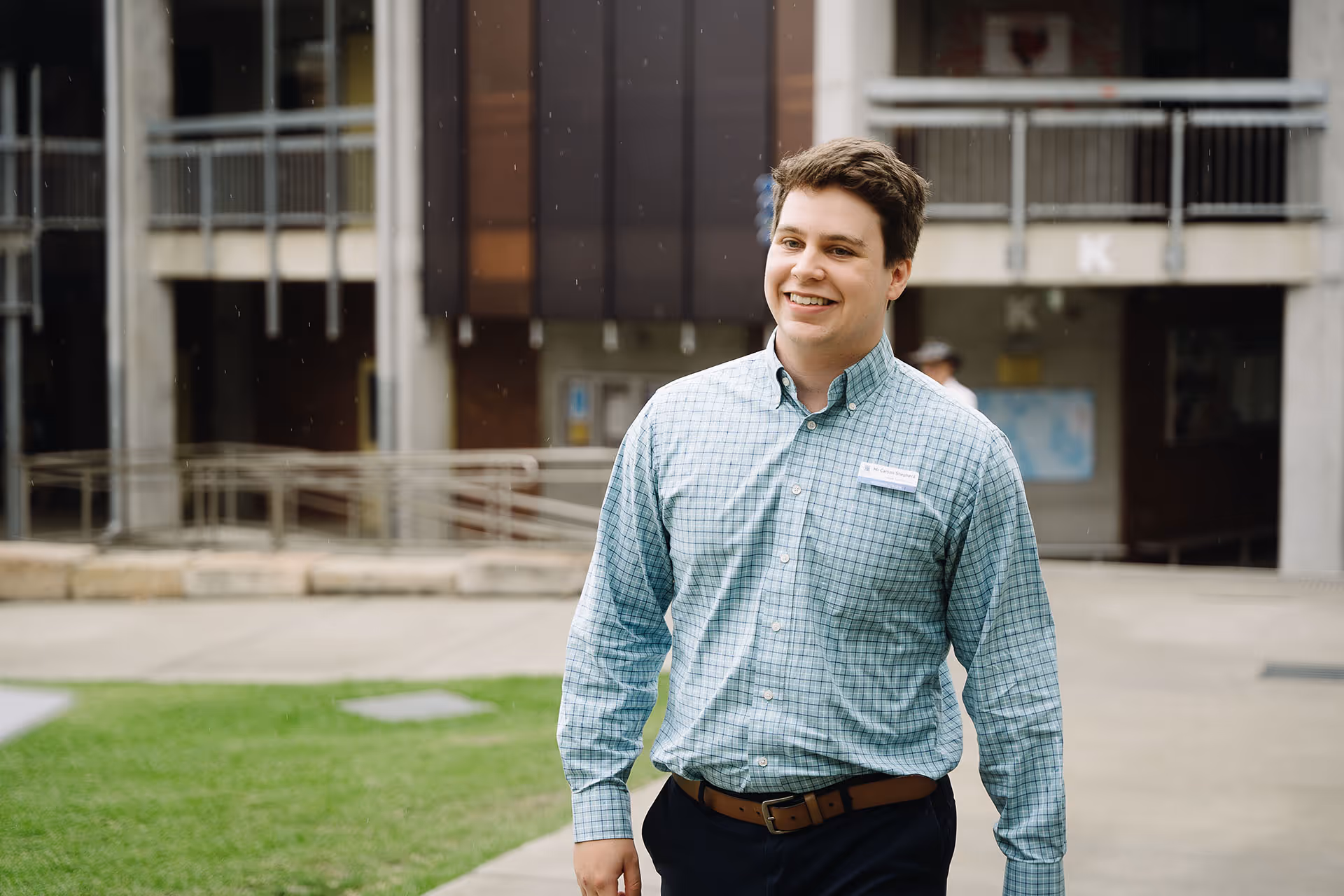 Smiling young man in a checked shirt walking outdoors with a building and green lawn in the background.