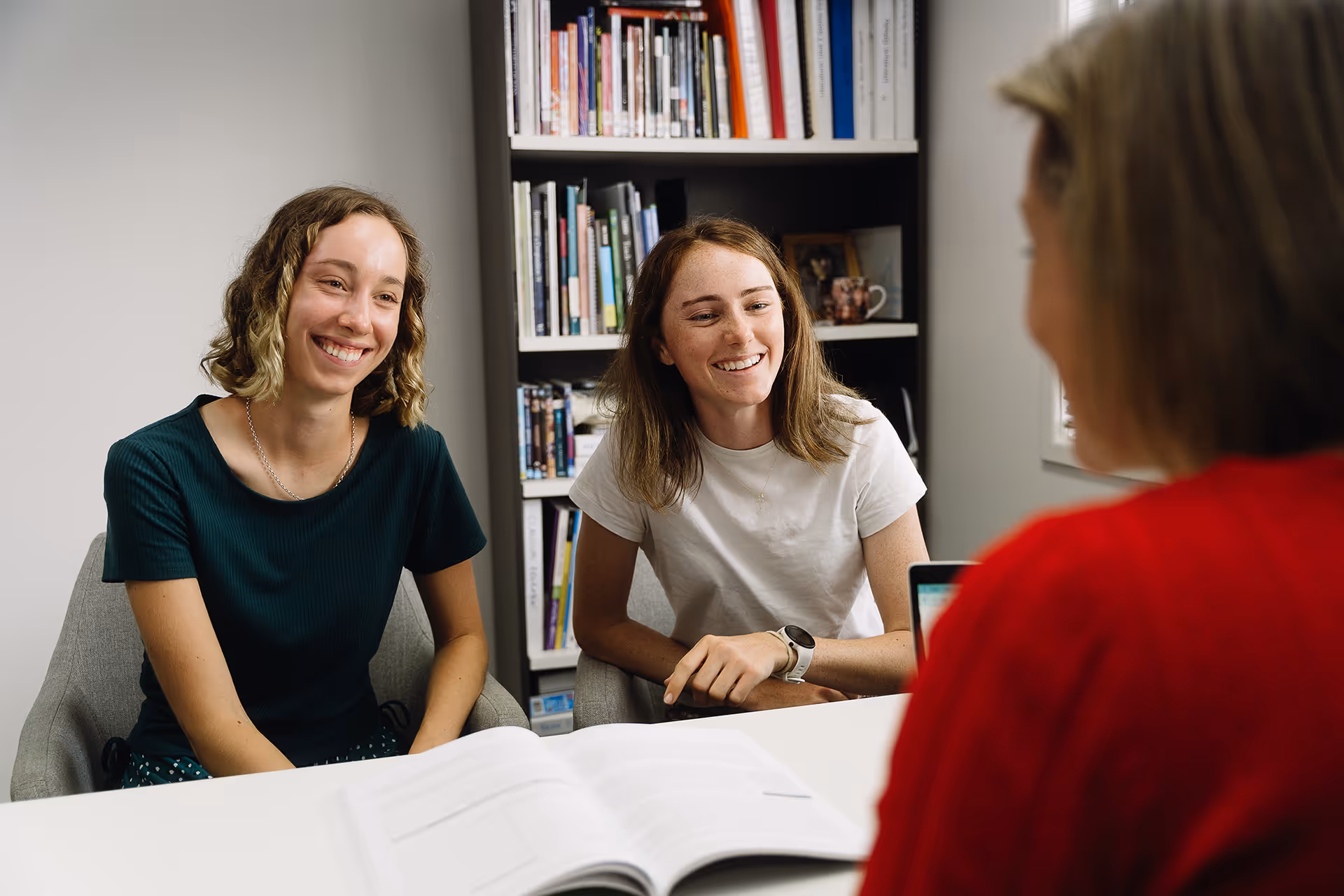 Two young women smiling and sitting at a table with an open book, talking to a person in a red shirt opposite them.