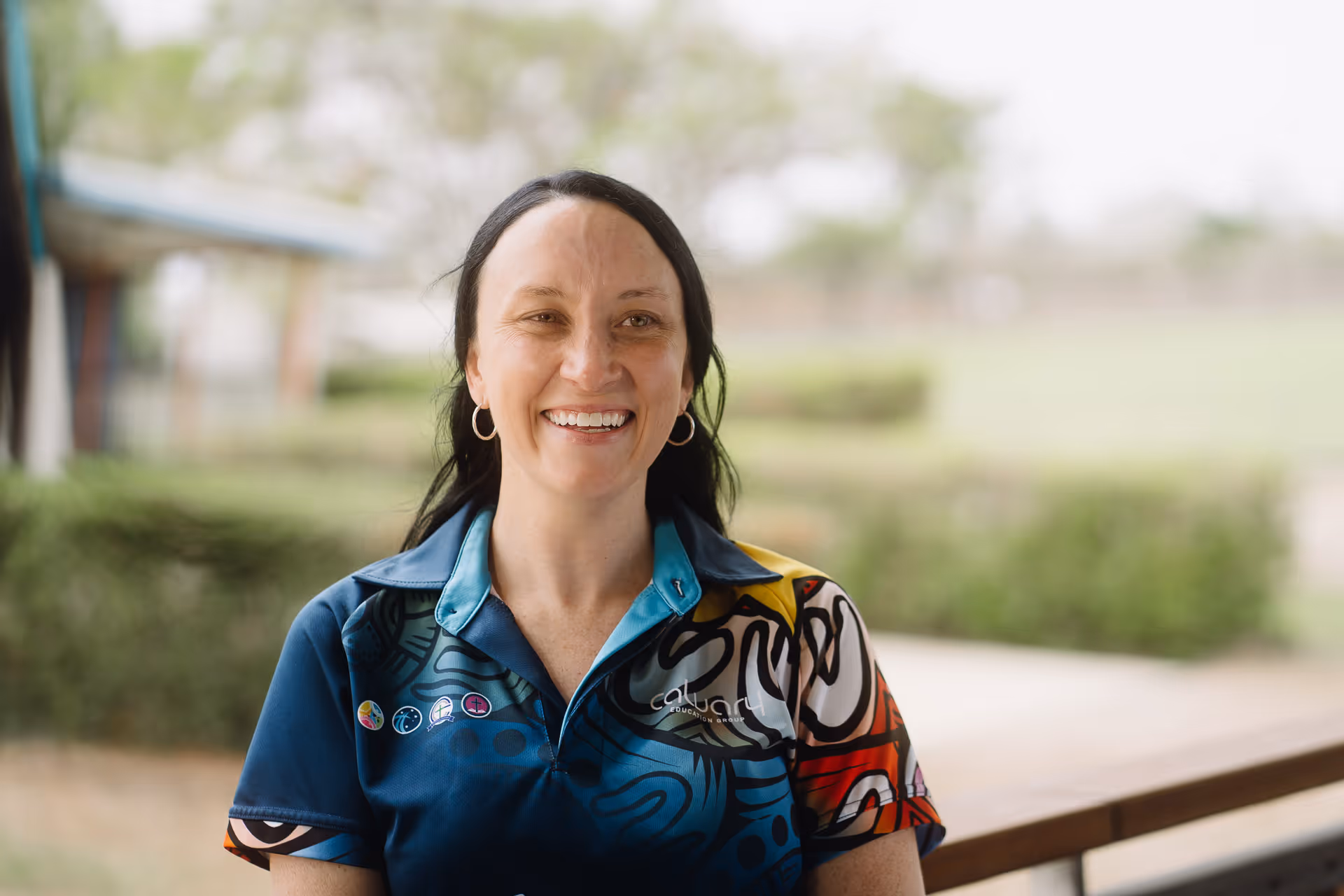 Smiling woman with dark hair and hoop earrings wearing a colorful Calvary Education Group shirt outdoors.