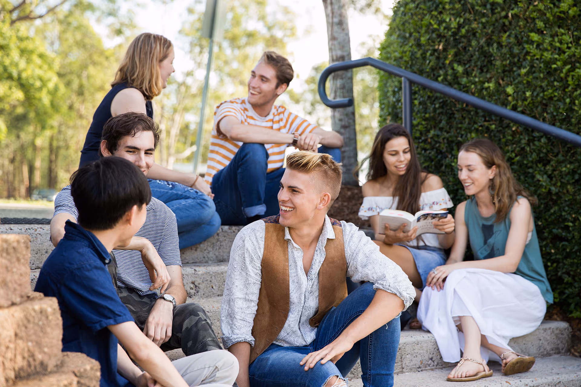 Group of six young adults sitting on outdoor steps chatting and smiling on a sunny day.