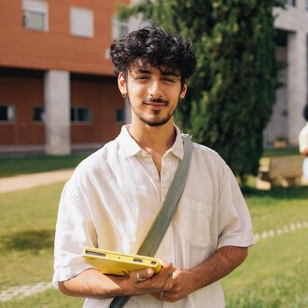 Smiling young man with curly hair wearing a white button-up shirt and gray shoulder bag holds a yellow laptop outdoors on a sunny day.