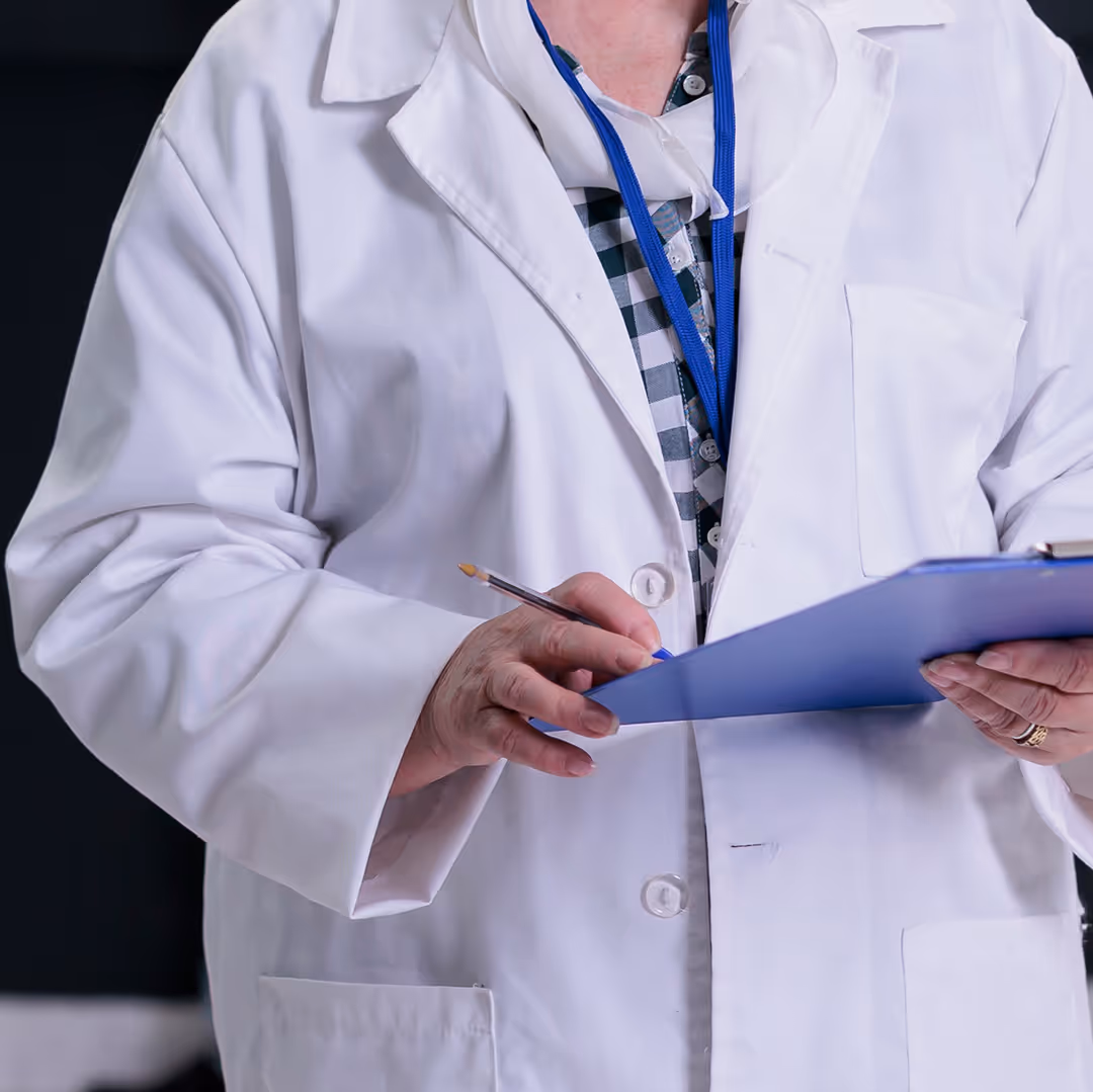 Person wearing a white lab coat and blue lanyard writing on a blue clipboard.