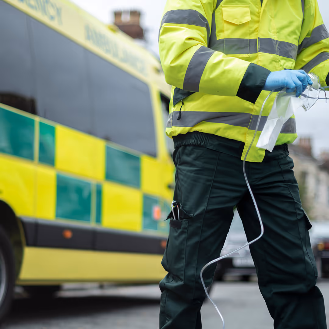 Paramedic in high-visibility uniform holding oxygen mask near an ambulance.