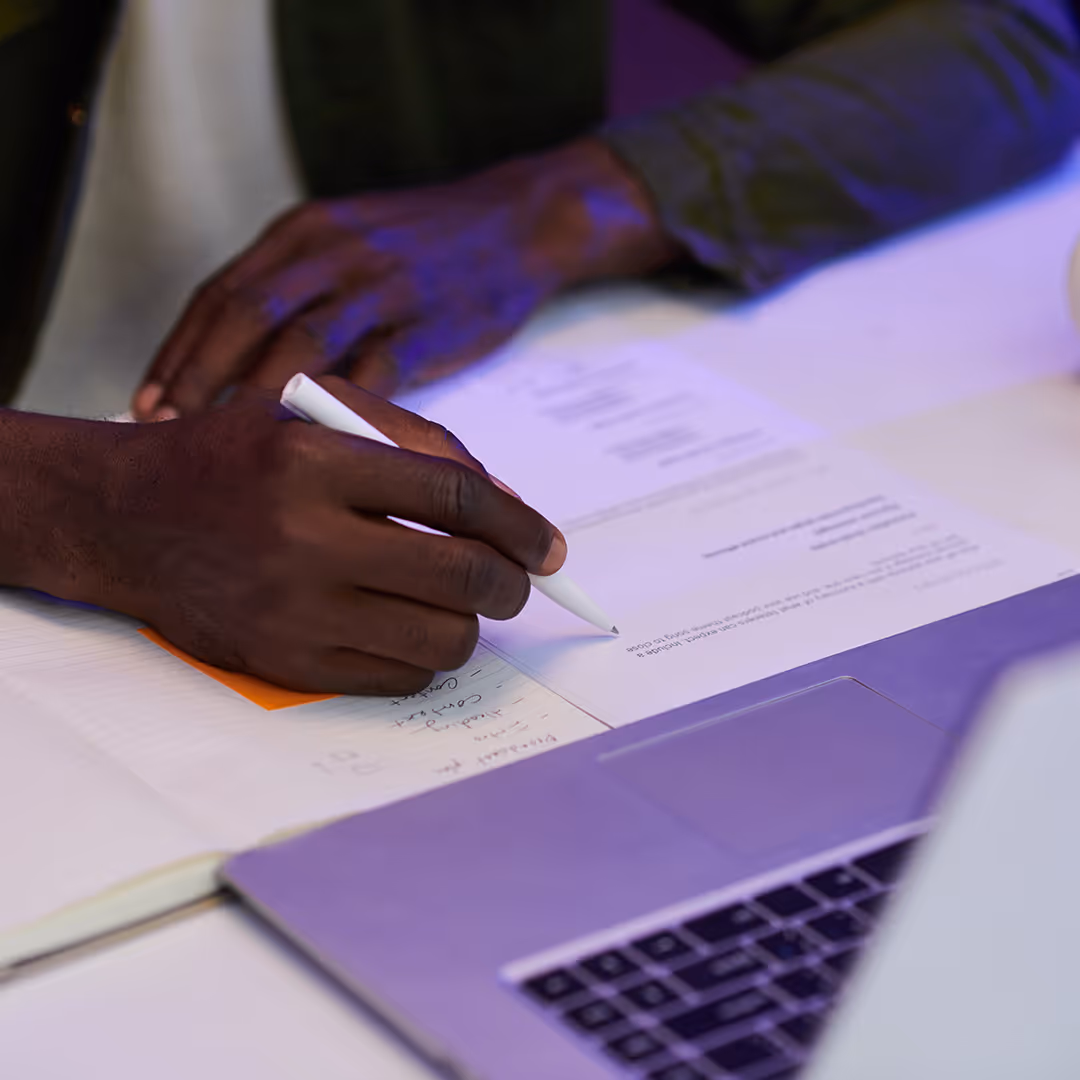 Person writing on printed documents with a white pen next to a laptop keyboard.