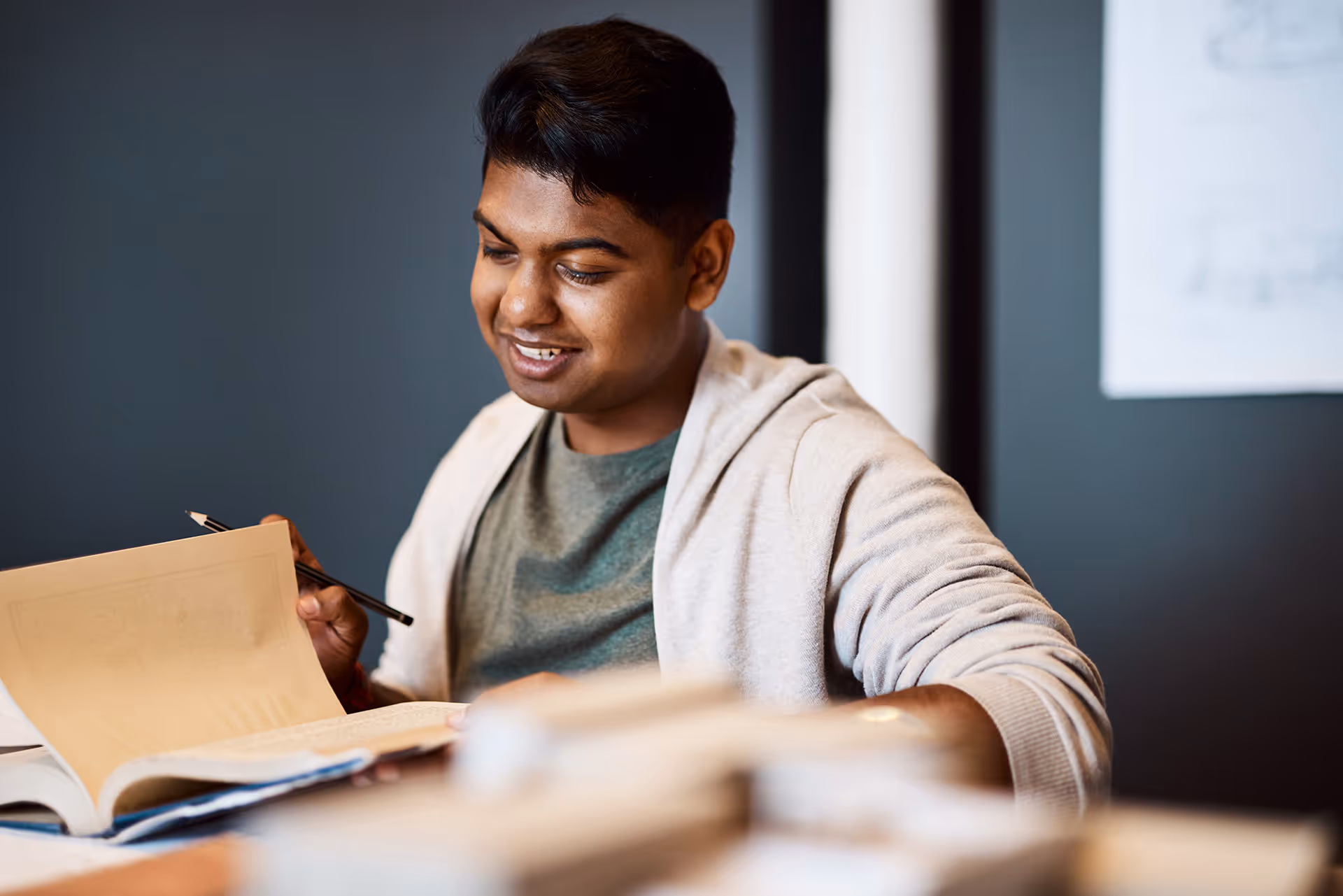 Smiling young man sitting at a table reading and holding a pencil with books around him.