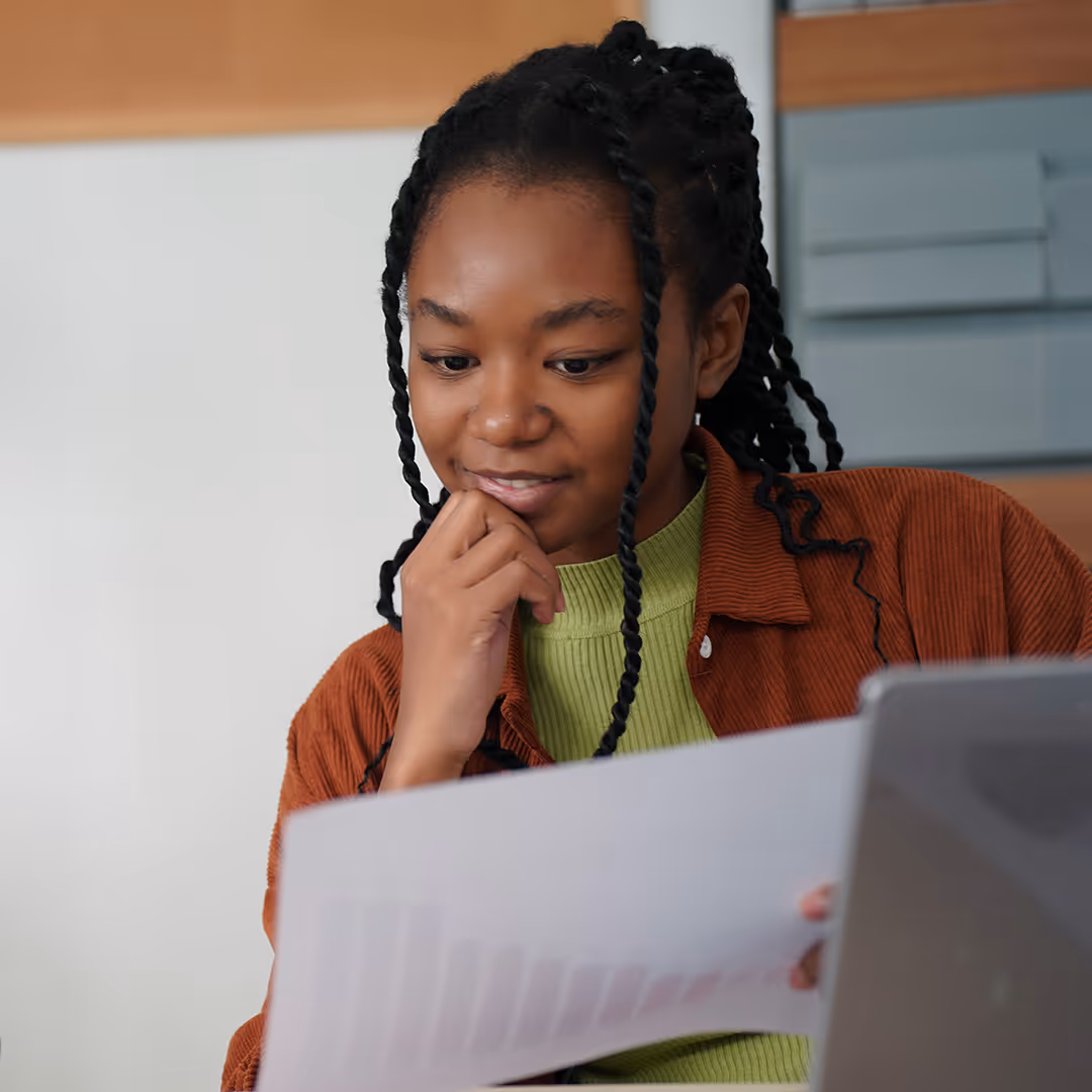 Young woman with braided hair reading a document while seated at a desk with a laptop.