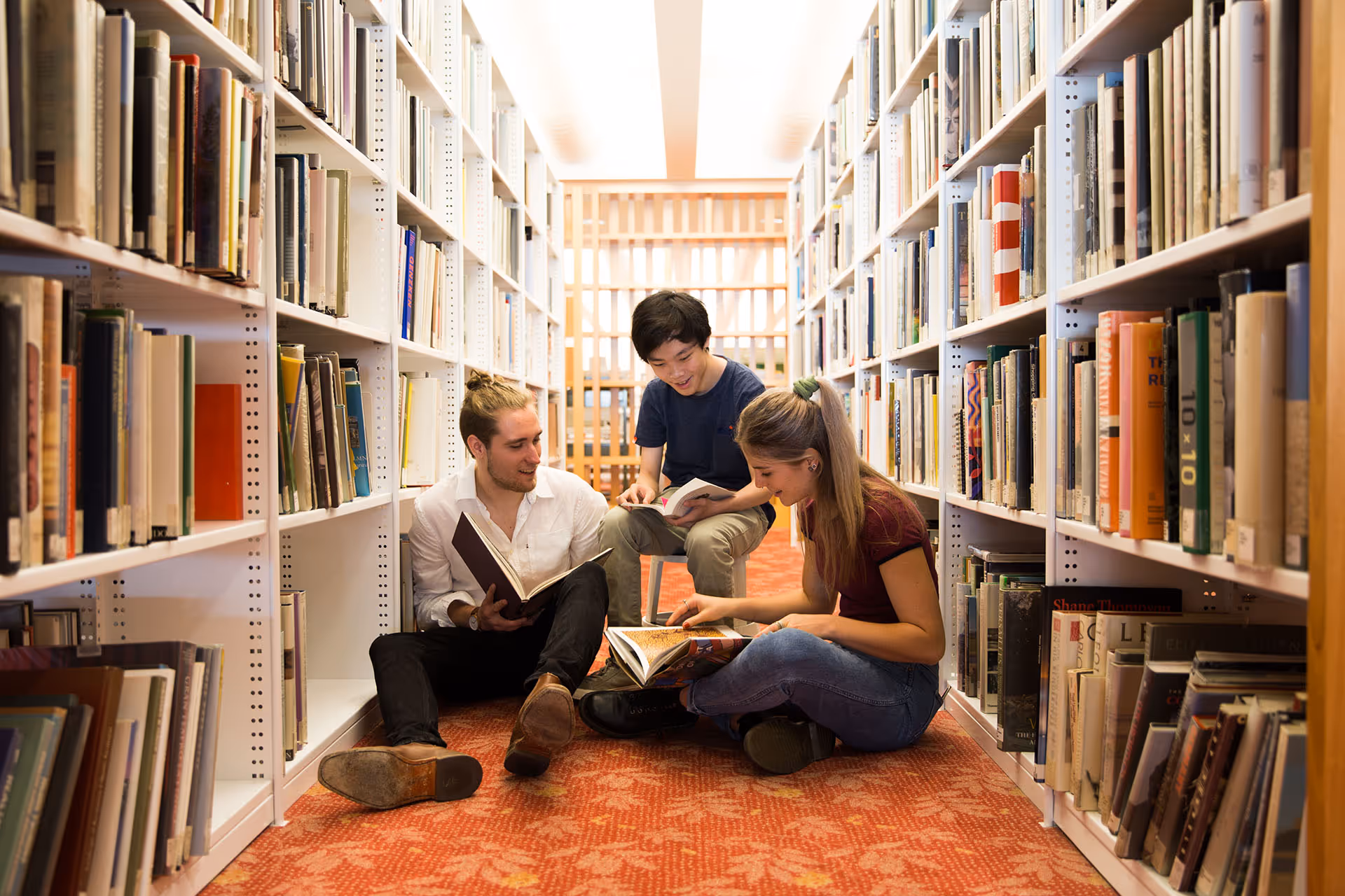 Three students sitting on a patterned carpeted floor between tall white bookshelves in a brightly lit library, reading books and smiling.