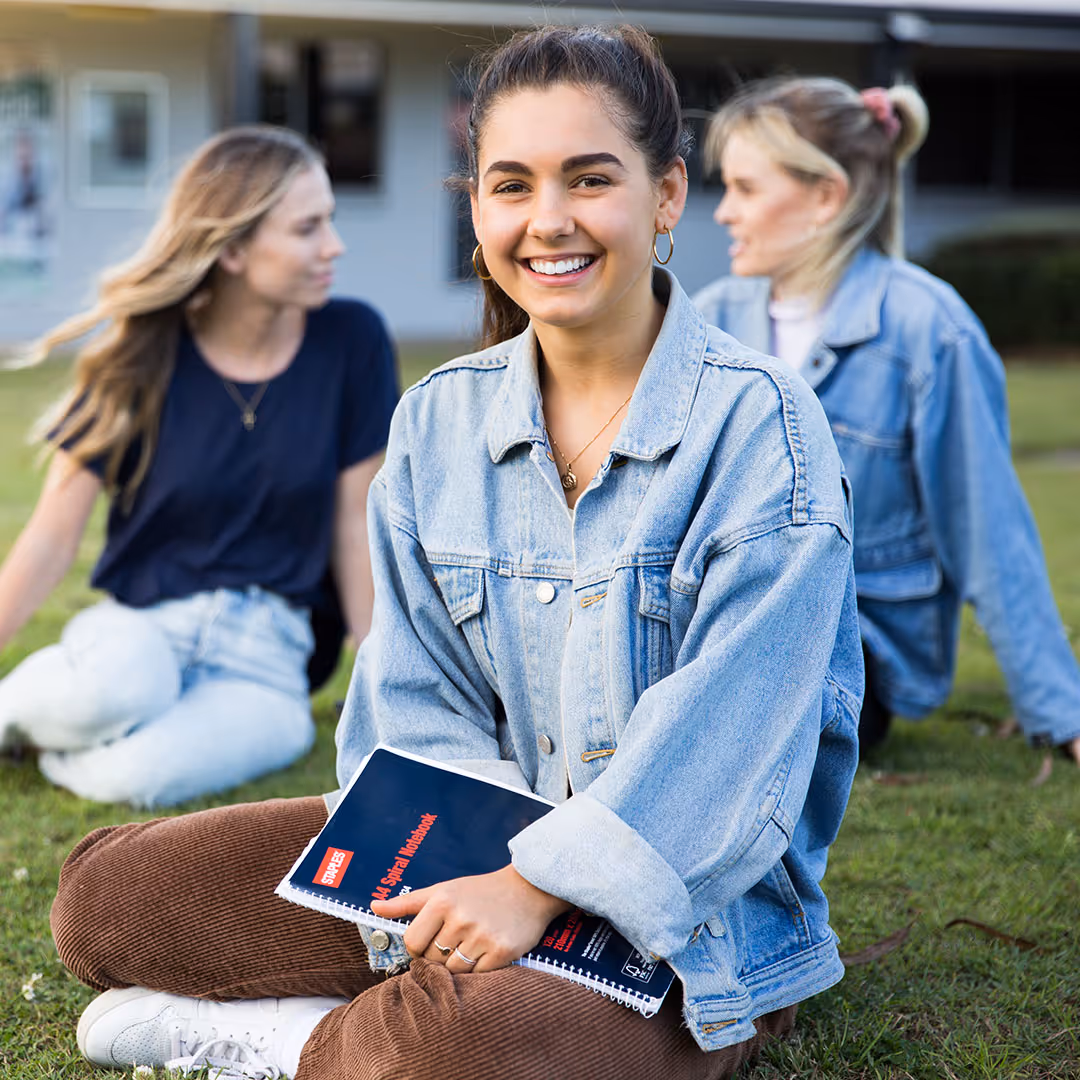 Smiling young woman in a denim jacket sitting cross-legged on grass holding a spiral notebook, with two other women in the background.