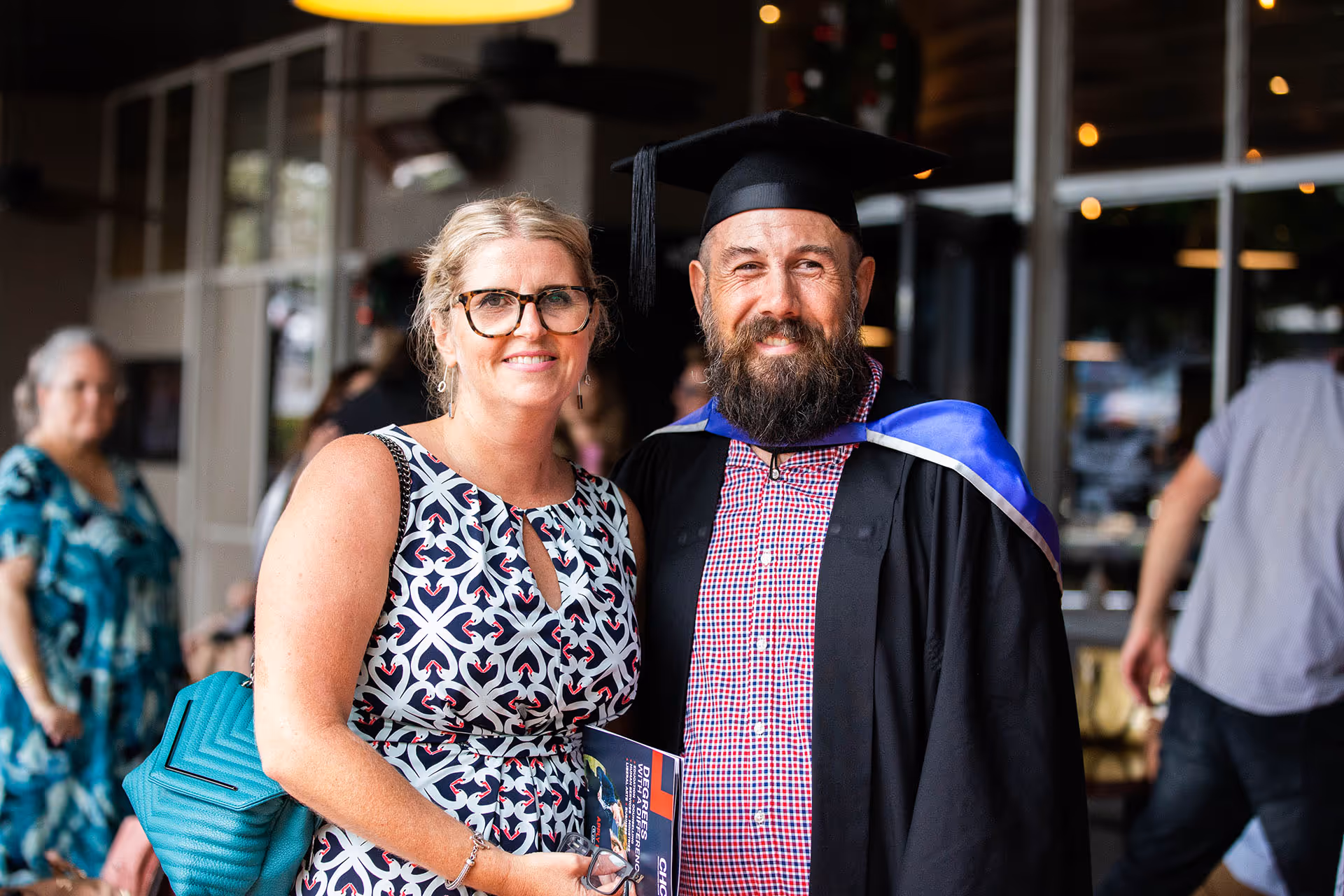 Smiling graduate in cap and gown standing next to a woman in a patterned dress inside a building.