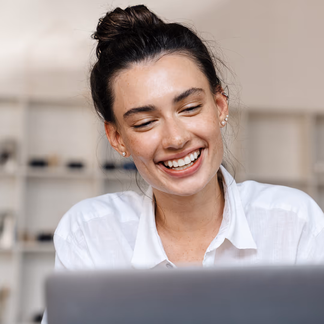 Smiling woman with dark hair tied up looking at a laptop screen.