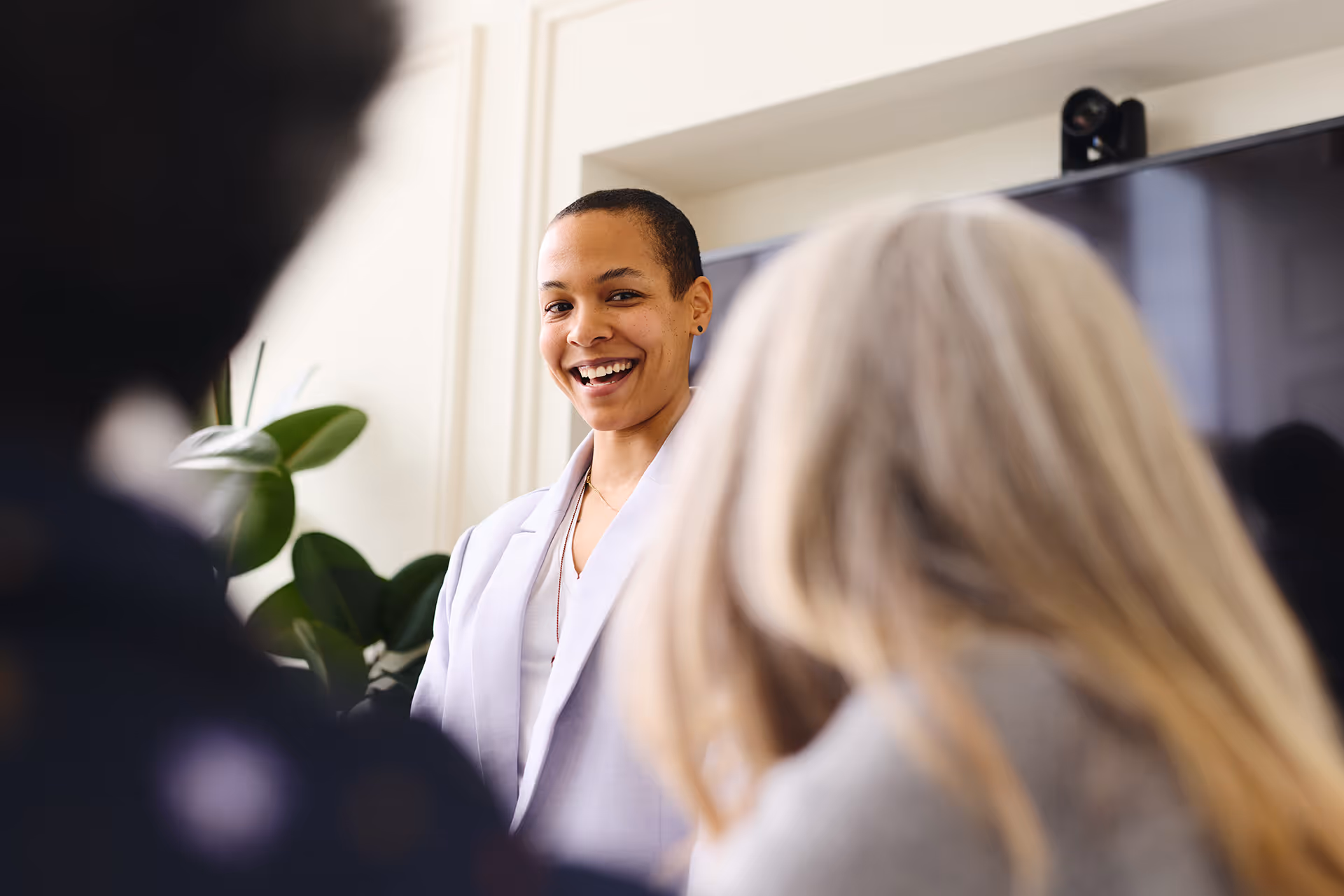 Smiling person in a white blazer speaking to a small audience in an indoor setting.